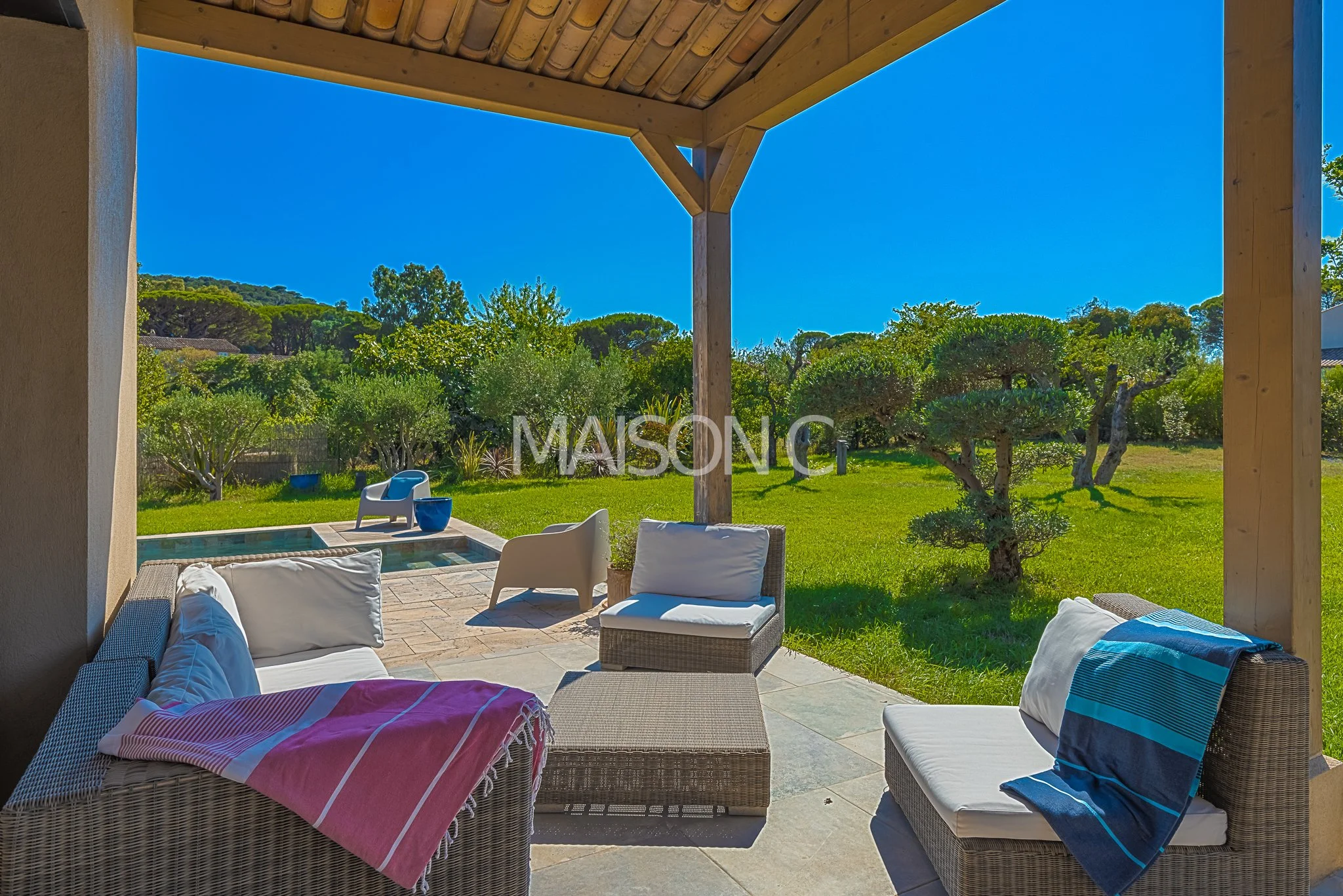 Terrasse avec mobilier de jardin sous un toit en bois, vue sur un jardin verdoyant avec arbres et un petit bassin d'eau, ciel bleu clair.
