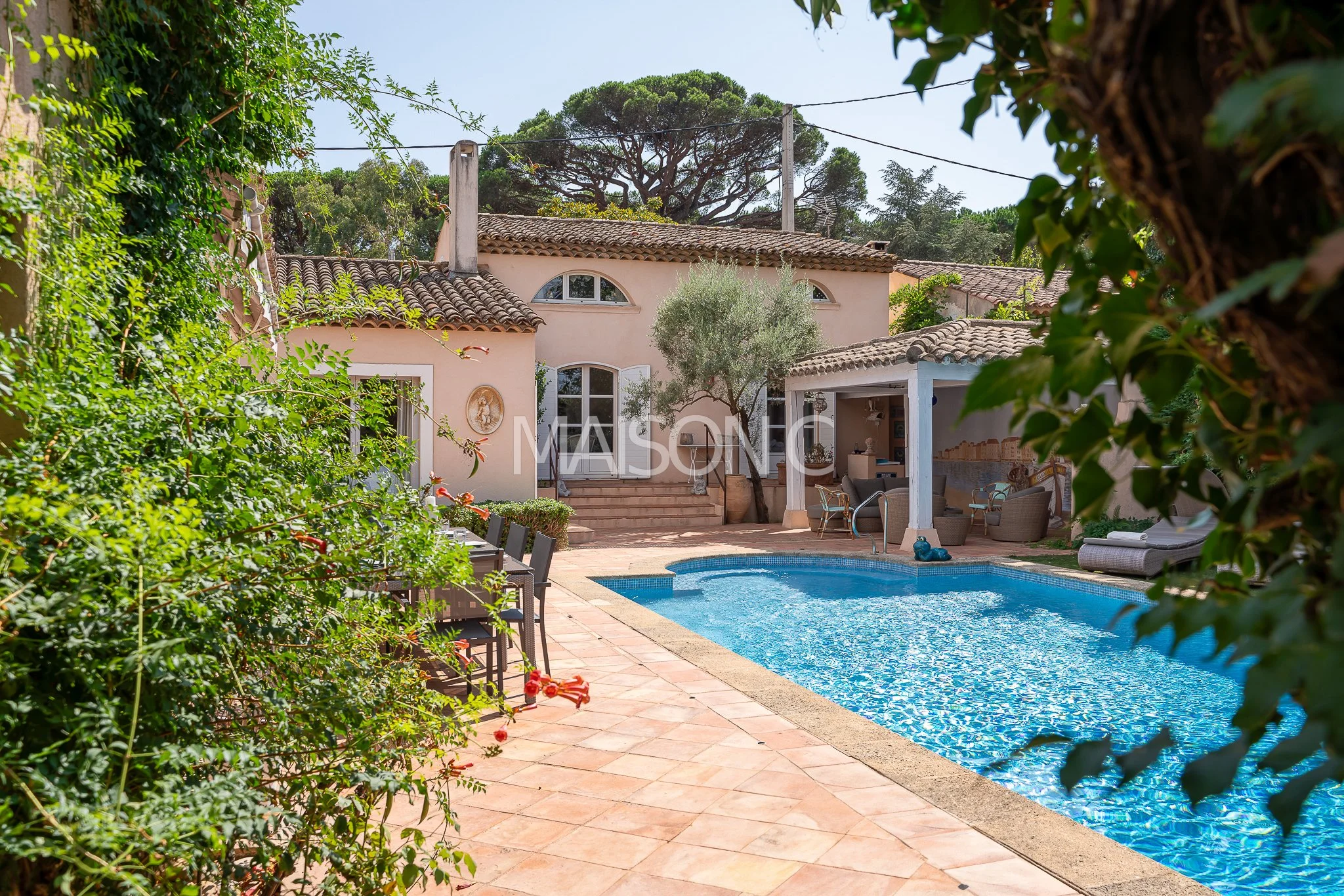 Maison de style méditerranéen avec piscine dans un jardin lush, mobilier de patio et arbres sous un ciel ensoleillé.