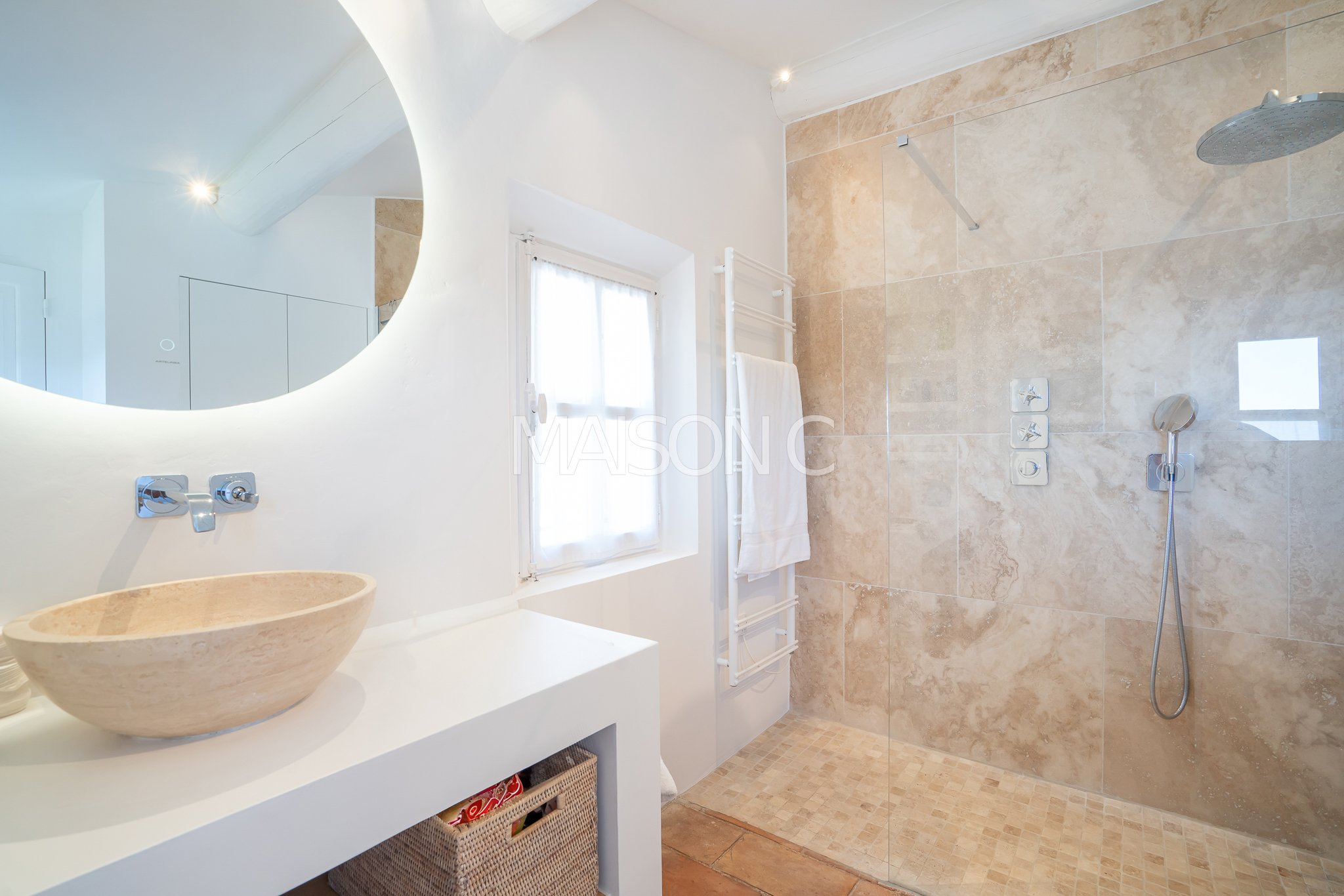 Salle de bain avec douche à l'italienne, grand miroir rond, vasque en pierre naturelle, fenêtres avec rideaux blancs, radiateur sèche-serviettes blanc, murs en pierre beige, sol en carrelage beige.