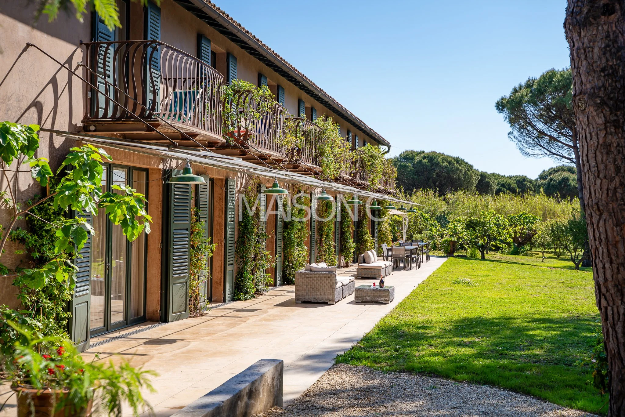 Facade d'une maison avec terrasse et salon extérieur, entourée de végétation et d'arbres, sous un ciel bleu clair.