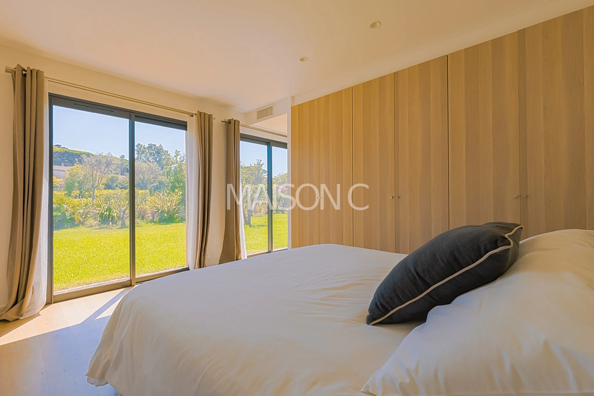 Chambre spacieuse avec vue sur un jardin, lit avec oreillers, murs en bois et fenêtres grandes laissant entrer la lumière naturelle.