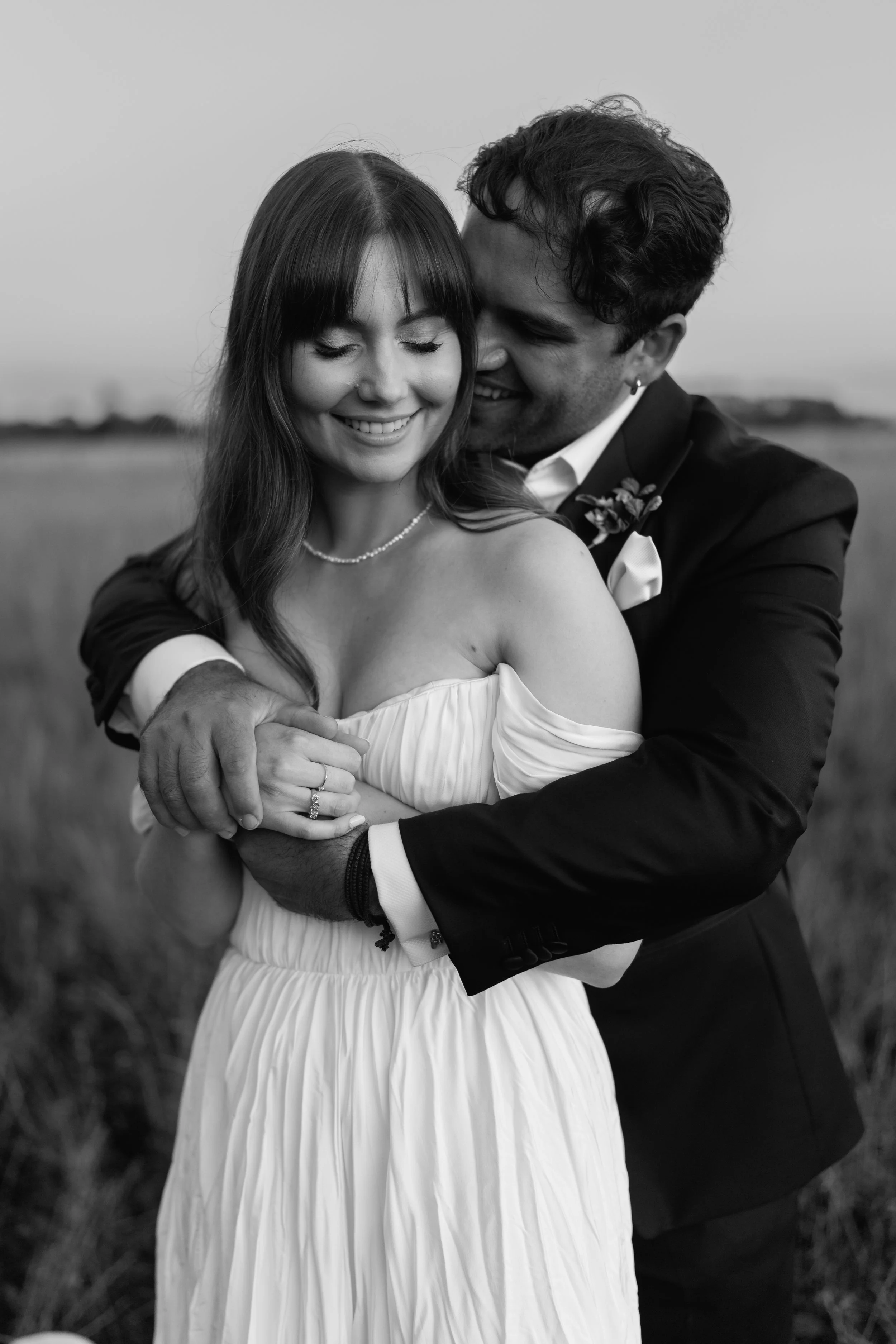 A black and white photo of a happy couple embracing outdoors. The woman is wearing a white off-shoulder dress and a pearl necklace. The man is in a dark suit and white shirt, holding her with both arms. They are smiling with eyes closed, in a field or open natural setting.