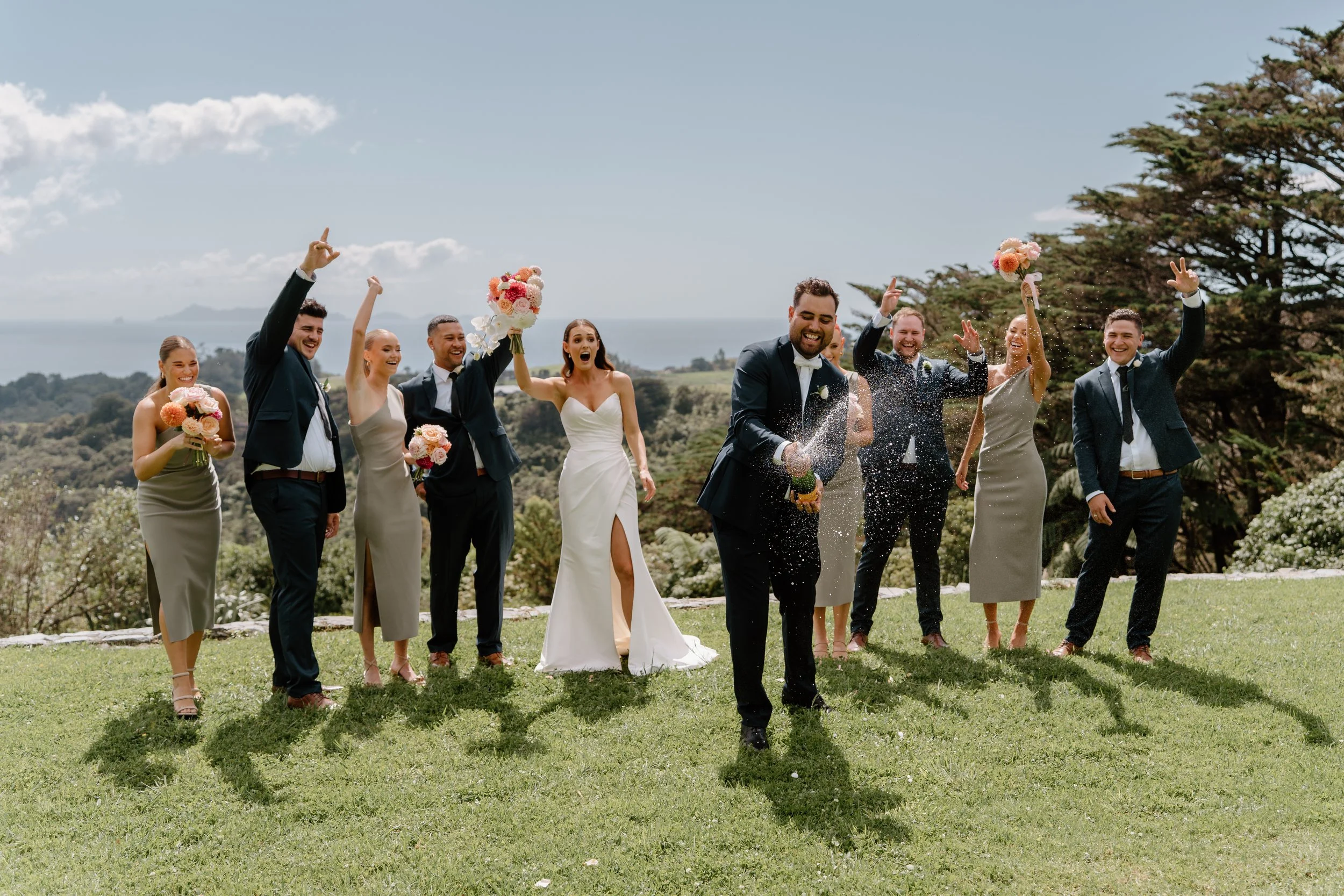 Celebrating wedding ceremony outdoors with a group of men and women dressed in formal attire, holding bouquets and cheering on a groom who is spraying champagne on a grassy area with trees and ocean in the background.