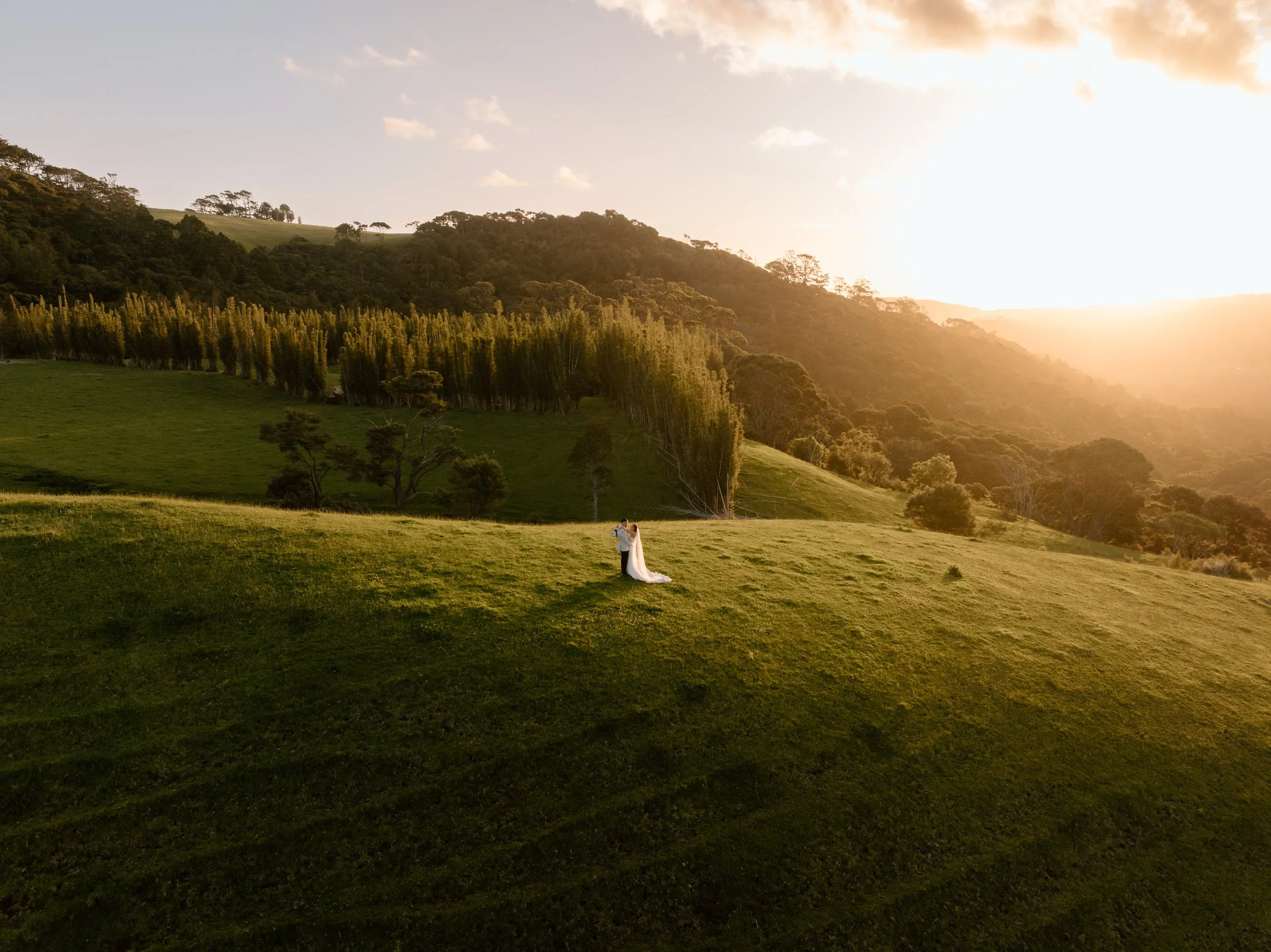 A bride and groom standing on a grassy hill during sunset with a scenic mountain landscape in the background.
