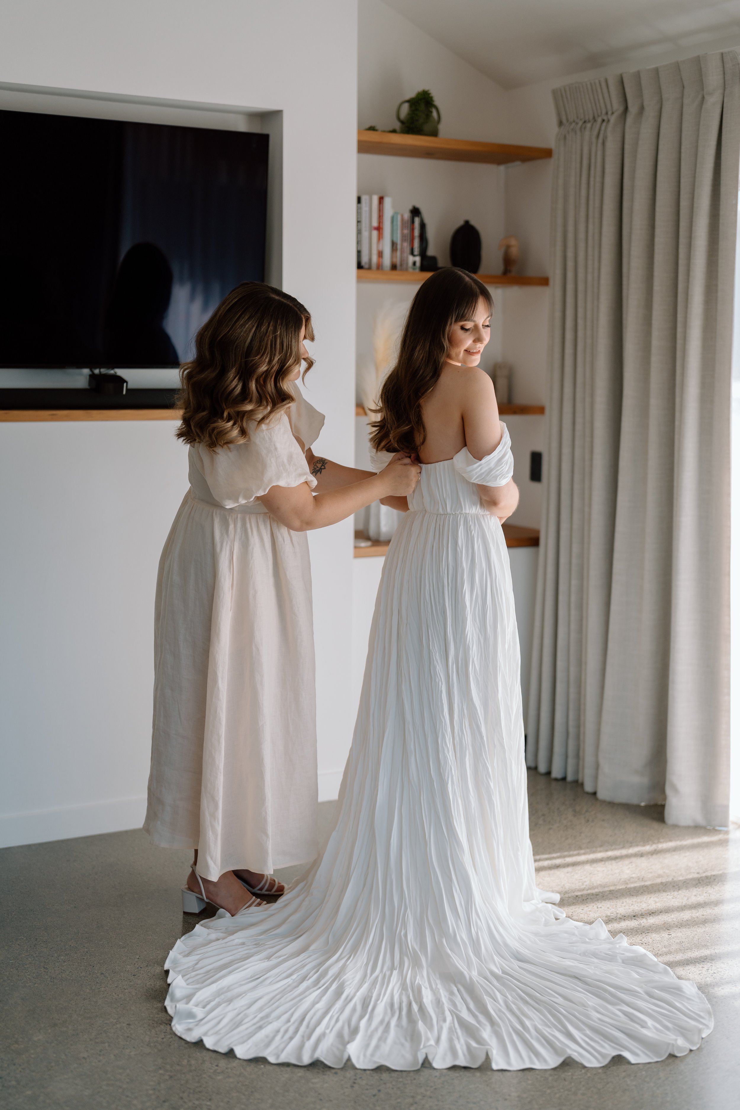 A woman in a white wedding dress being helped with her dress by another woman in a beige dress, in a modern room with beige curtains and wooden shelves.