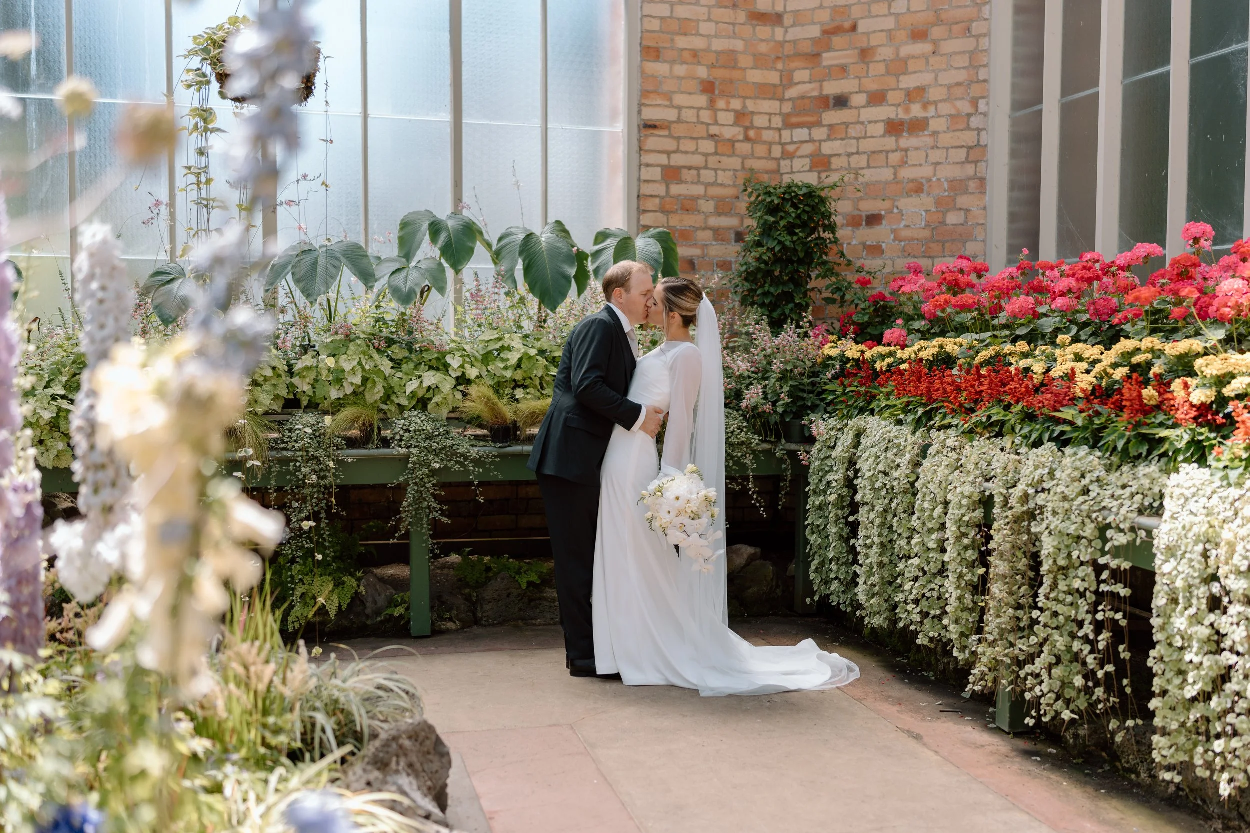 A bride and groom sharing a kiss in a greenhouse filled with colorful flowers, with the bride holding a bouquet of white flowers.