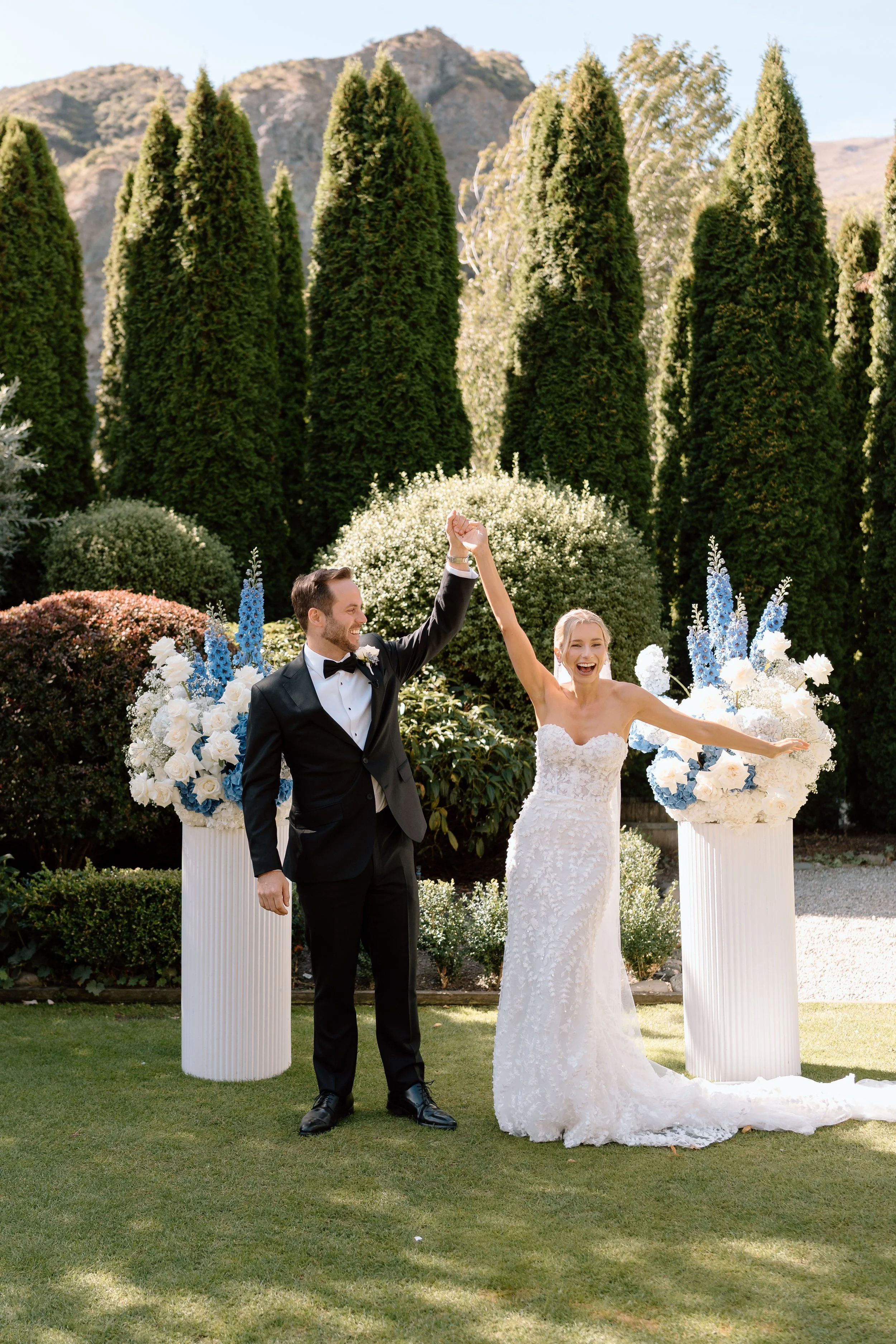 A newly married couple celebrating outdoors, holding hands with arms raised in victory. The groom is wearing a black suit with a bow tie, and the bride is in a white wedding dress, both smiling. They are surrounded by large flower arrangements on white pedestals and lush green trees with mountains in the background.