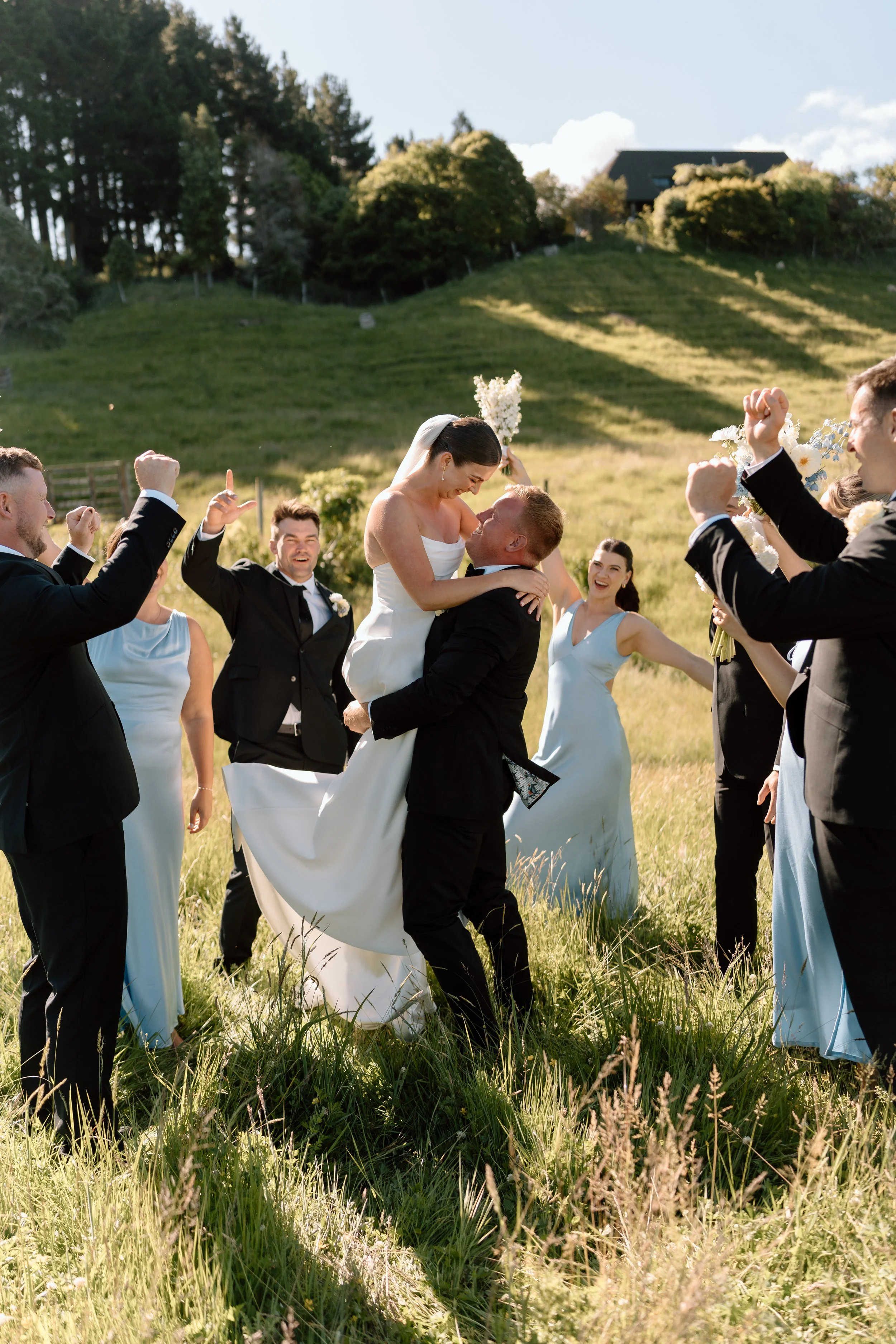 A wedding celebration outdoors with a bride and groom. The groom lifts the bride as friends and family cheer around them in a grassy field with trees and a house in the background.