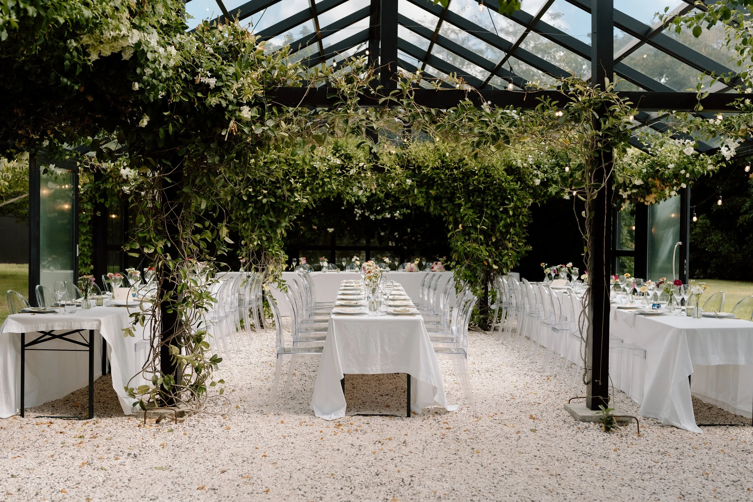 Elegant outdoor wedding reception setup with long white tables, transparent chairs, floral centerpieces, and a vine-covered pergola under a glass roof.