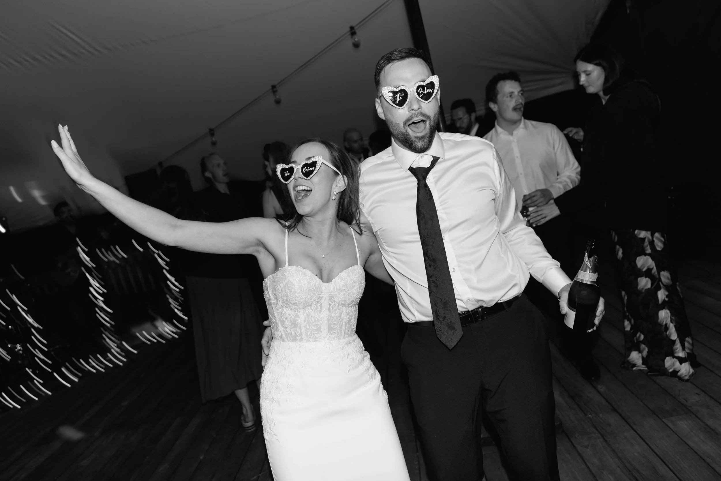 Two people, a woman in a wedding dress and a man in a white shirt and tie, wearing heart-shaped glasses with the words "The Bride" and "The Groom" written on them, celebrating at a wedding reception.