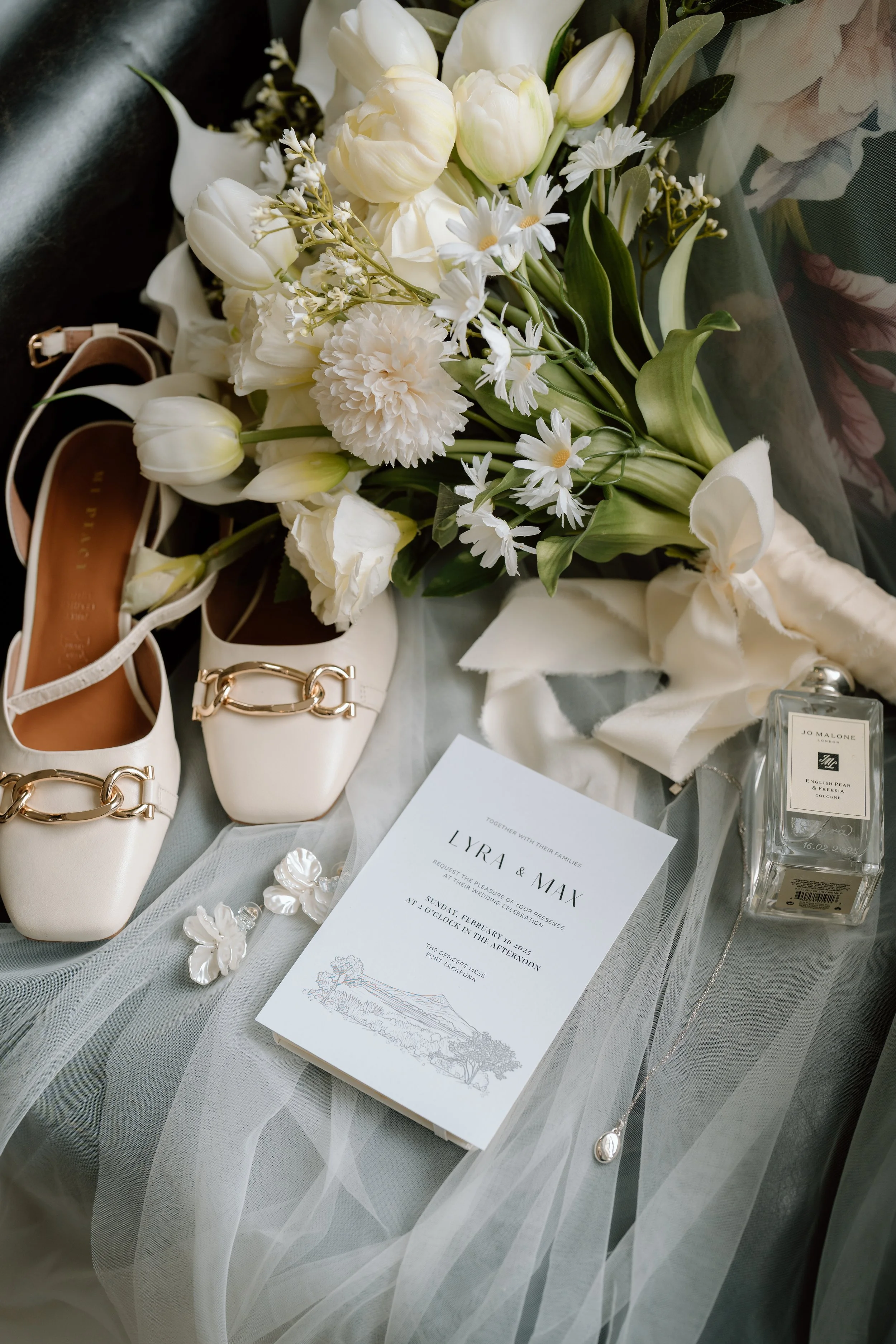 A wedding bouquet of white flowers, white shoes with gold chain accents, a wedding invitation, a bottle of Jo Malone cologne, and a pair of pearl earrings arranged on a soft fabric surface.