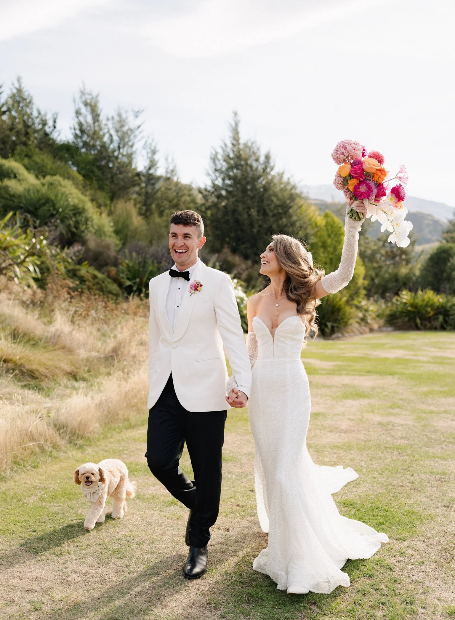 A newlywed couple walking hand in hand outdoors, smiling. The bride is holding a large colorful bouquet of flowers in her raised hand, and a small dog is walking beside them. The bride is wearing a white lace wedding gown, and the groom is in a white tuxedo jacket with a black bow tie.