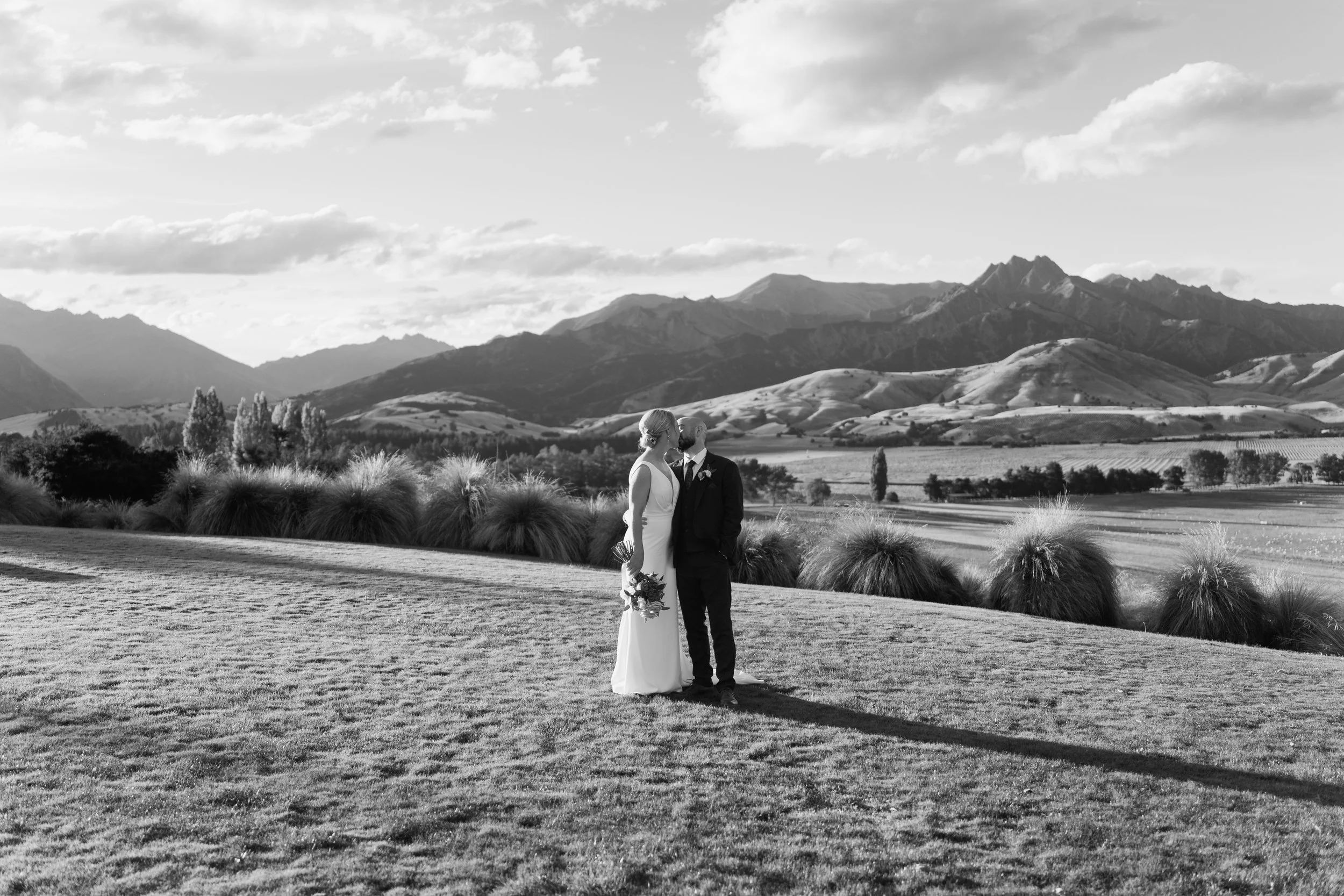 A bride and groom standing on grass in a scenic outdoor landscape with mountains and rolling hills in the background, black and white photograph.
