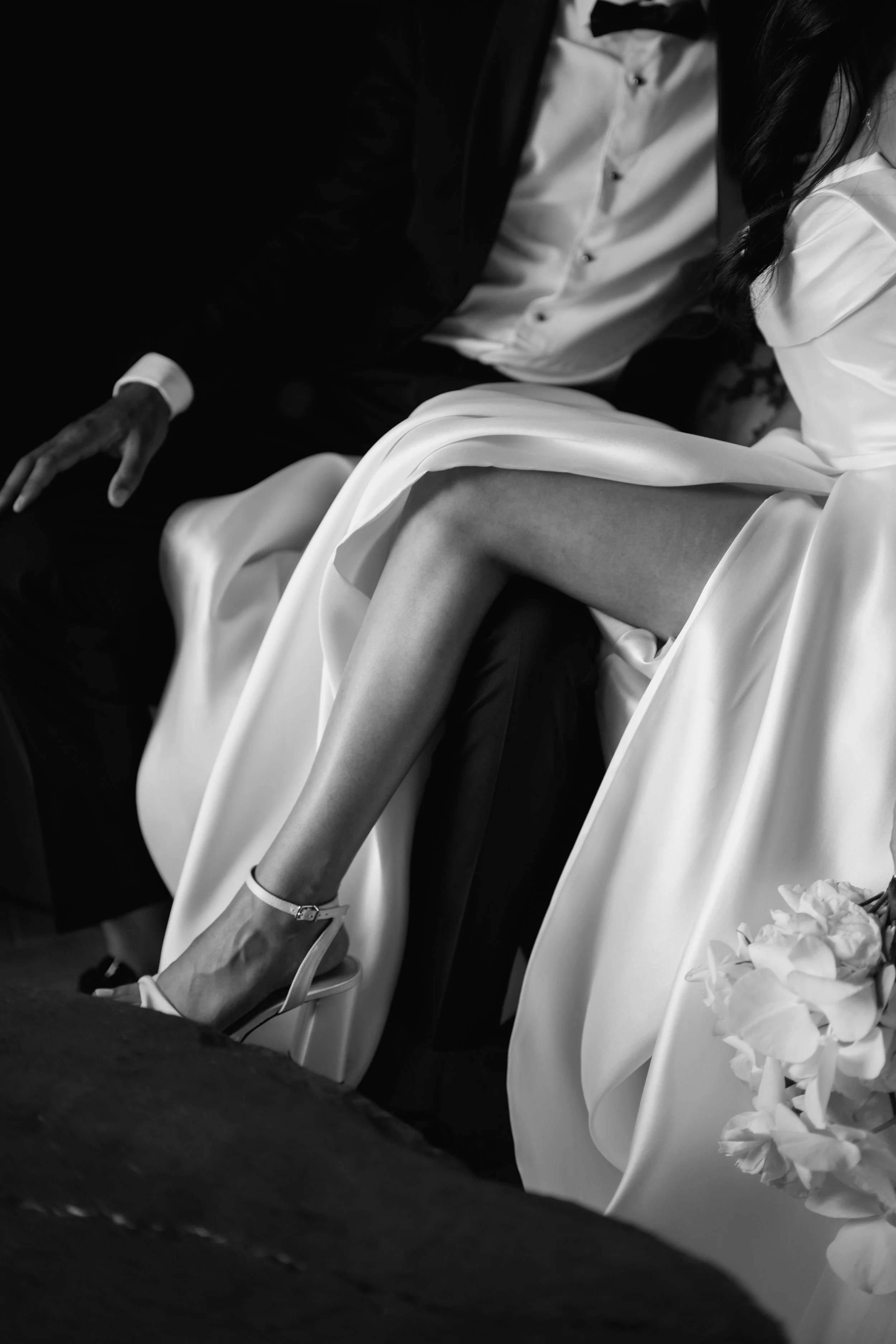 Close-up of a bride's leg in a wedding dress, paired with a high-heeled sandal, sitting beside someone dressed in a tuxedo.