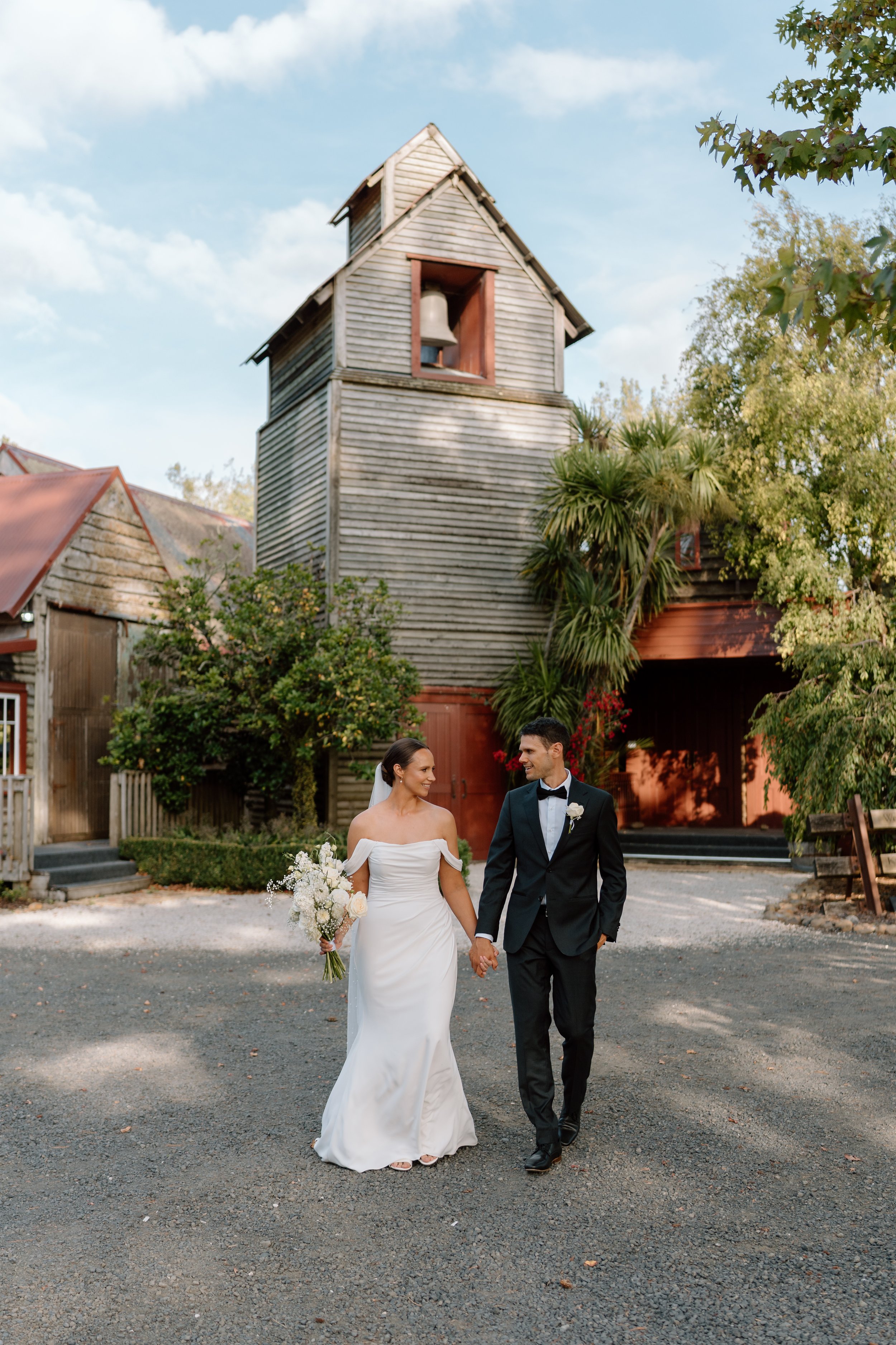 A bride and groom walking hand in hand outside, with the bride holding a bouquet of white flowers, in front of an old wooden building with a bell tower.