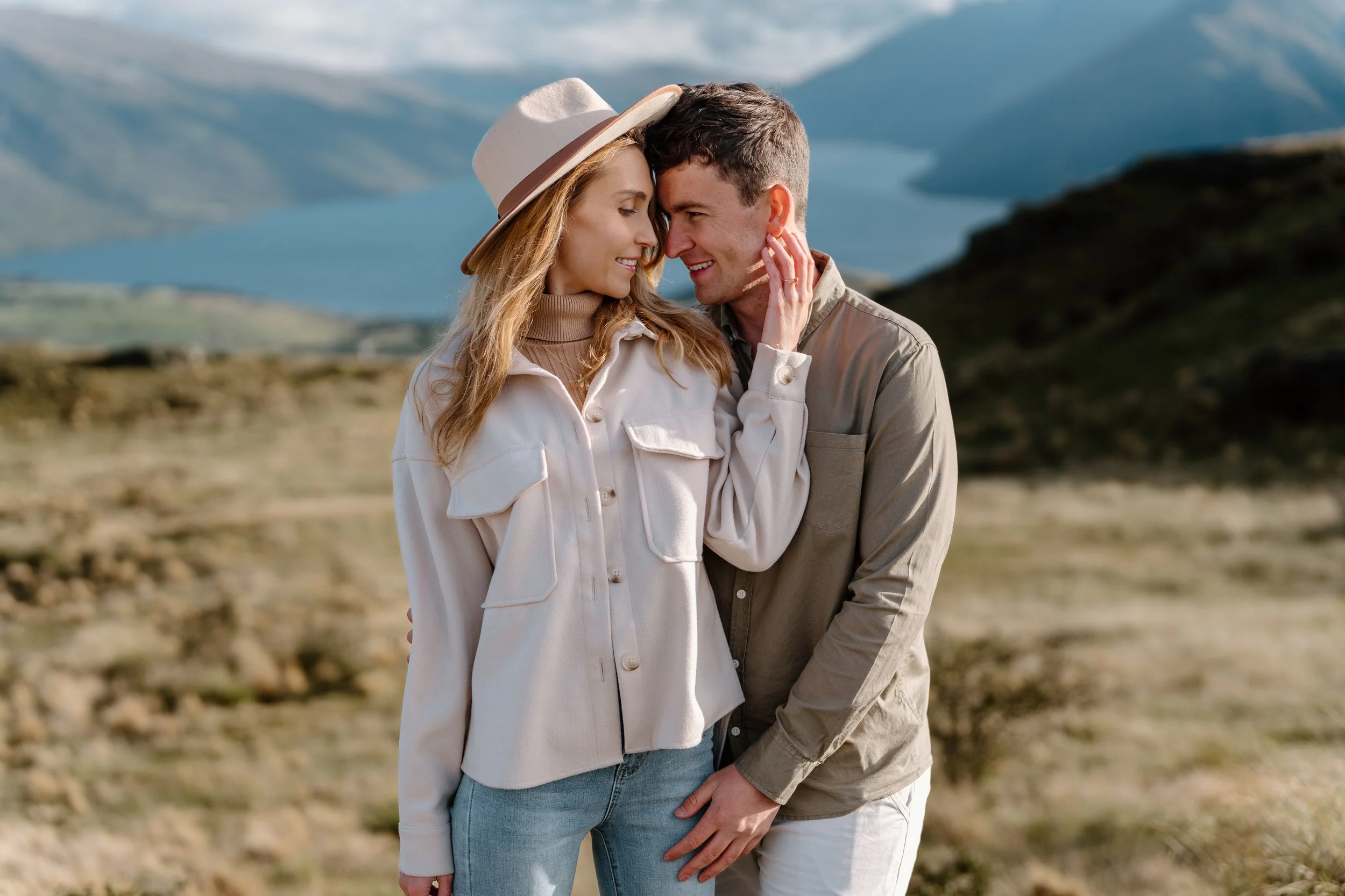 A couple standing close together outdoors in a scenic landscape with mountains and a lake in the background, sharing an intimate moment.