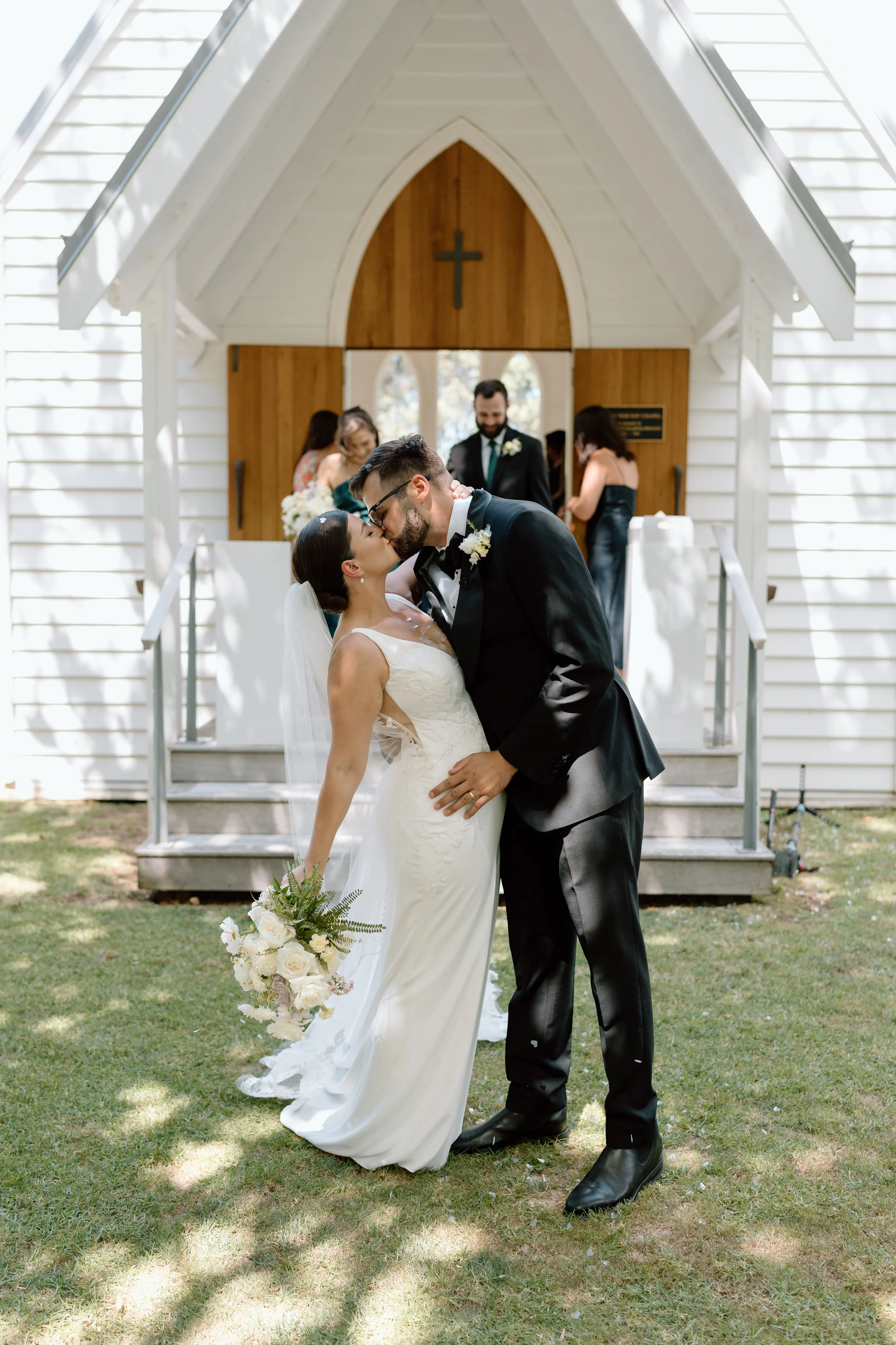 A newlywed couple kissing outside a small white chapel, with the bride holding a bouquet of white flowers and the groom in a black tuxedo.