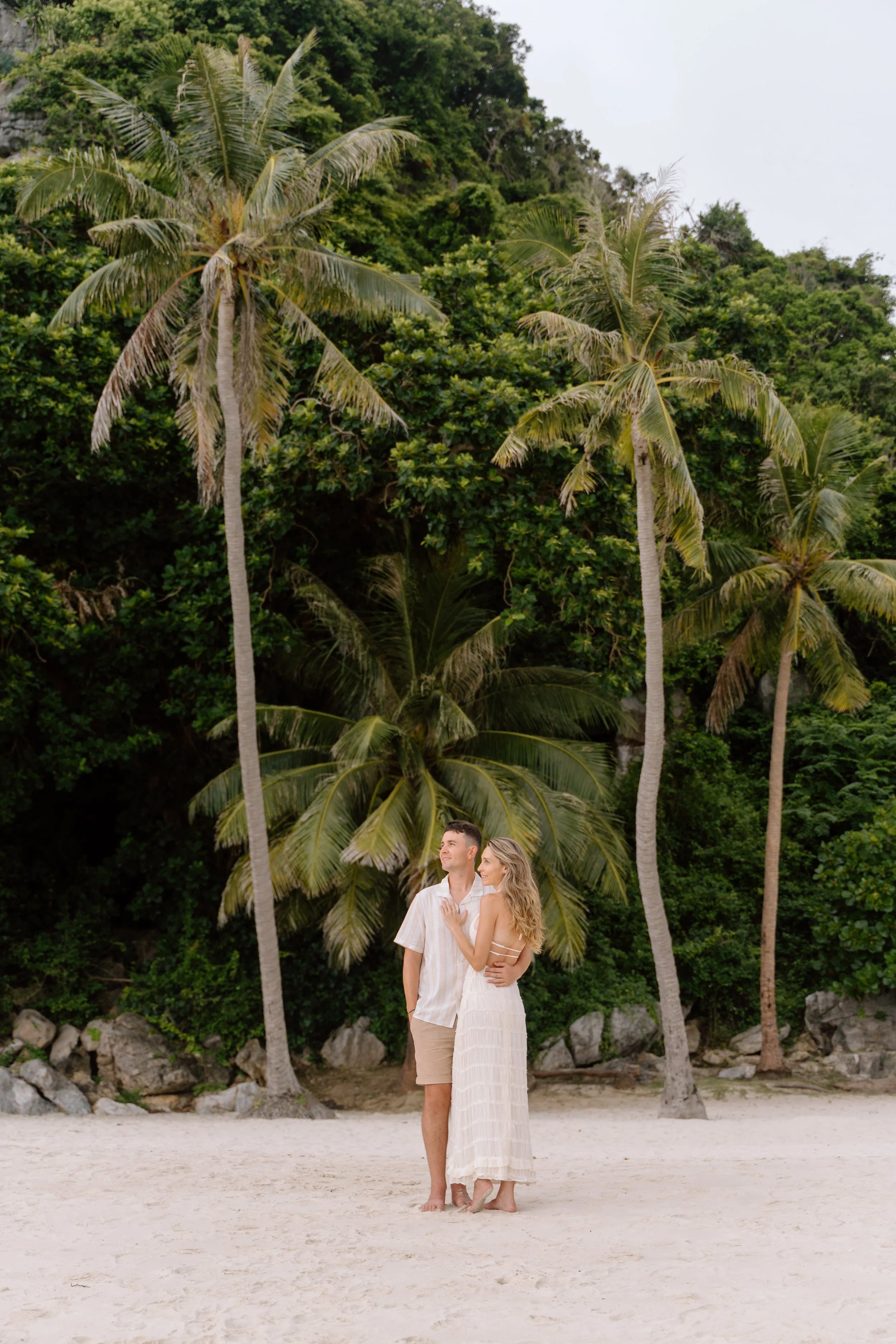 A couple standing on a sandy beach with lush green trees and tall palm trees in the background.