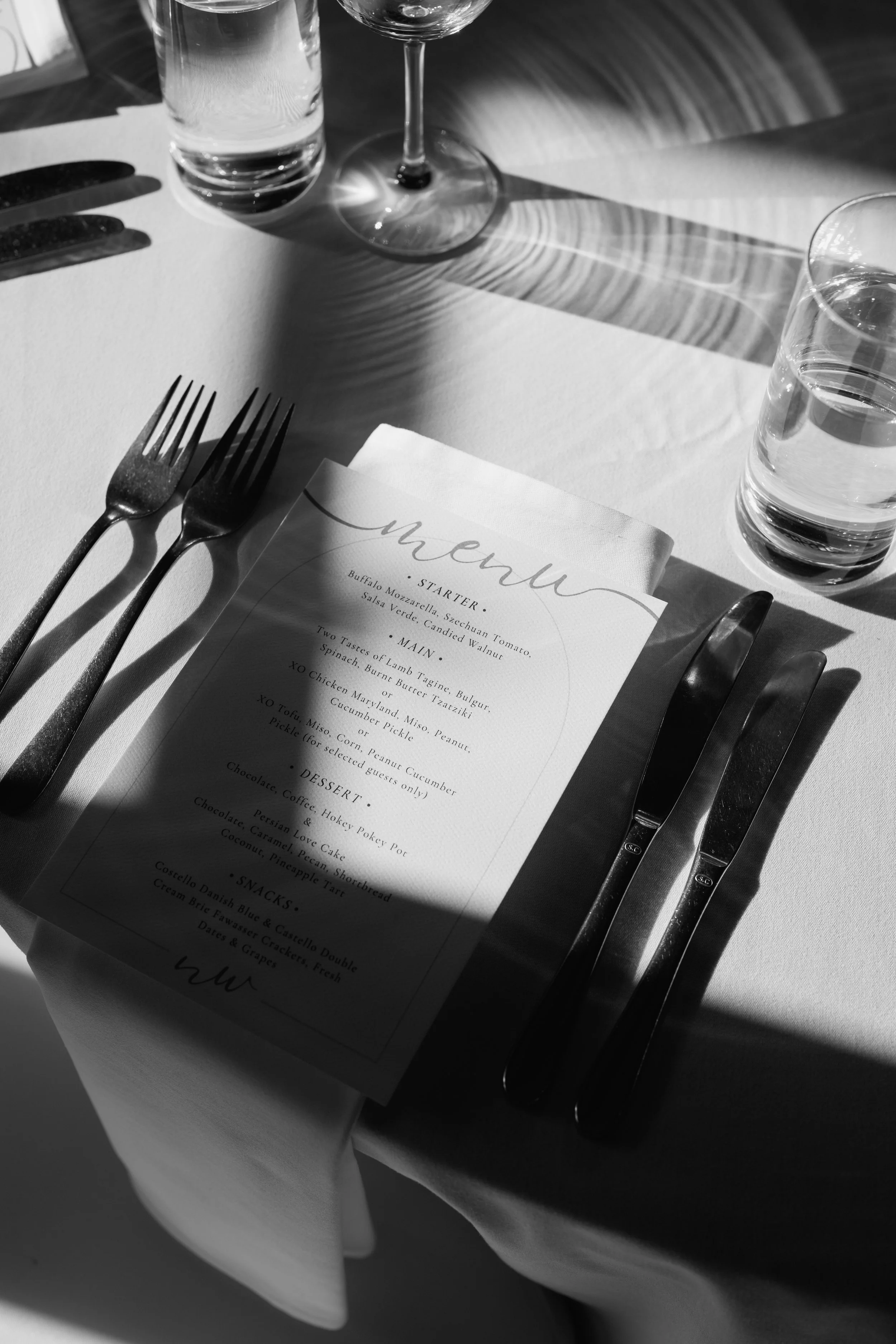 A black and white photo of a dining table setting with a menu, two forks on the left, and a knife and spoon on the right, with two glasses of water and a wine glass. Sunlight creates shadows on the table.