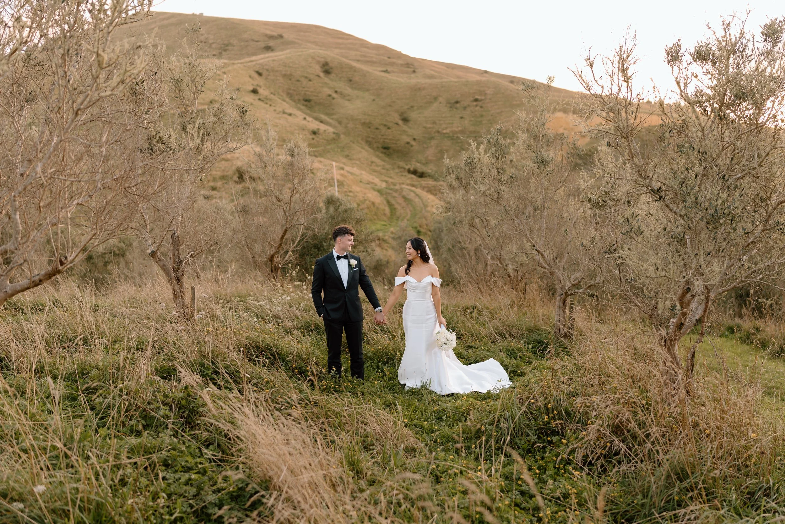 A bride and groom holding hands in a grassy, hilly landscape with sparse, leafless trees. The groom is wearing a black tuxedo, and the bride is in a white off-the-shoulder wedding dress, holding a bouquet of white flowers. The scenery looks like a natural, outdoor wedding setting.