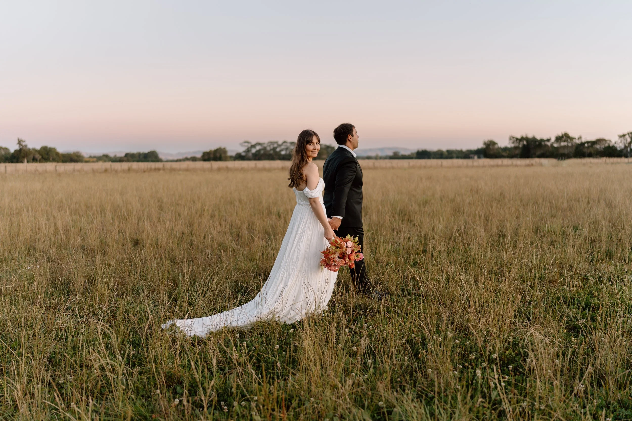Bride and groom holding hands in a grassy field at sunset, with the bride holding a bouquet of pink flowers, wearing a white wedding dress, and the groom in a black suit.