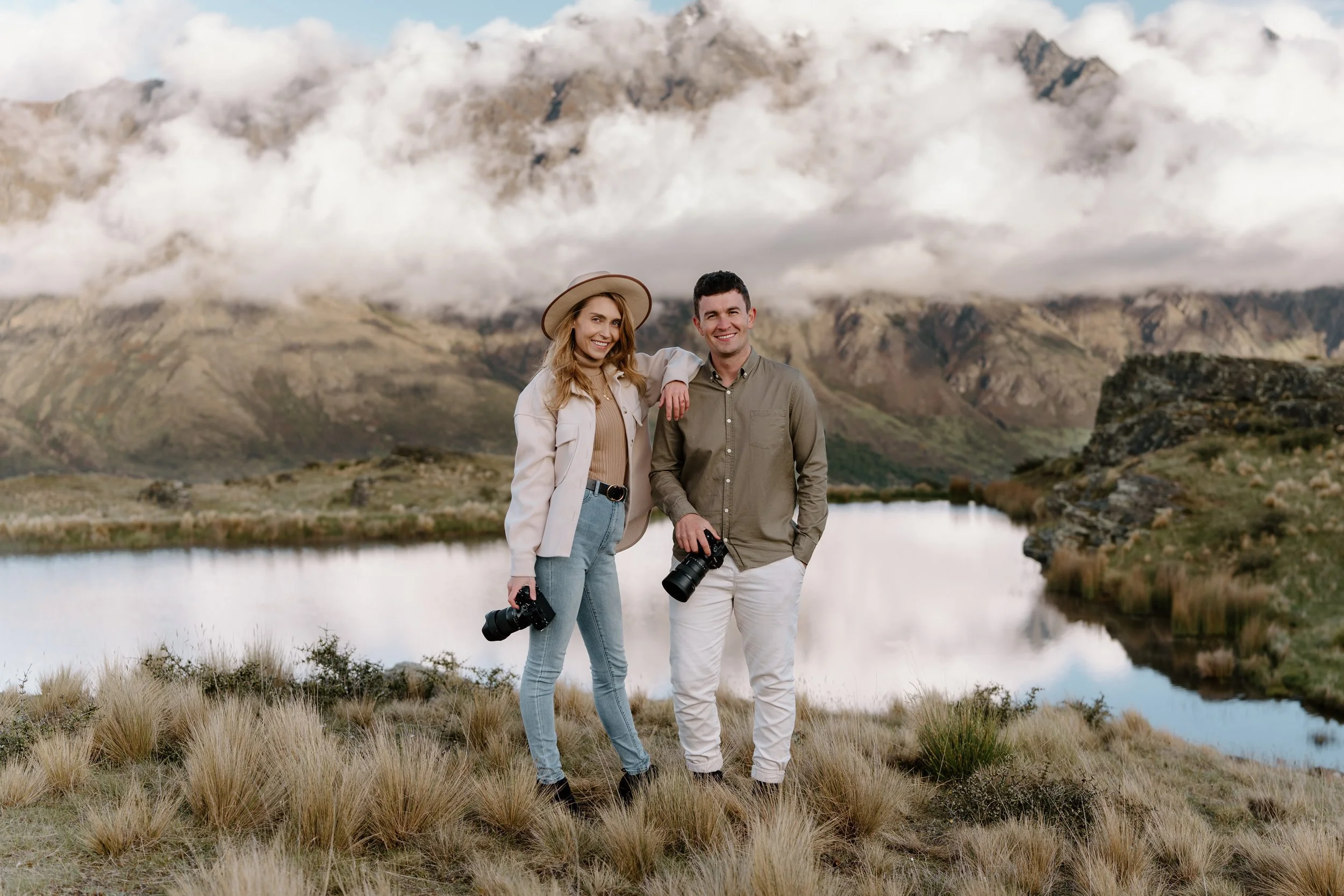 A smiling woman and man standing outdoors near a lake with mountains and clouds in the background, each holding a camera.