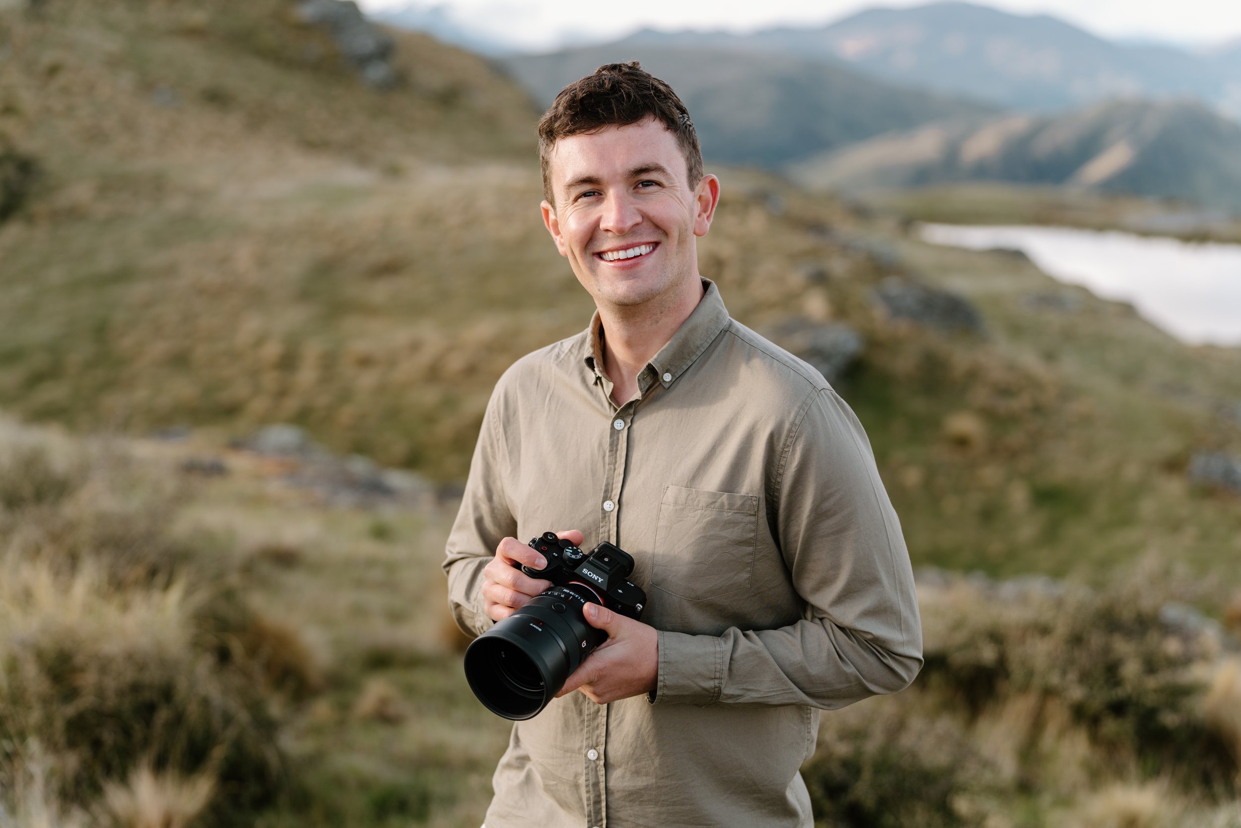 A smiling man holding a Sony camera outdoors in a grassy, mountainous landscape.
