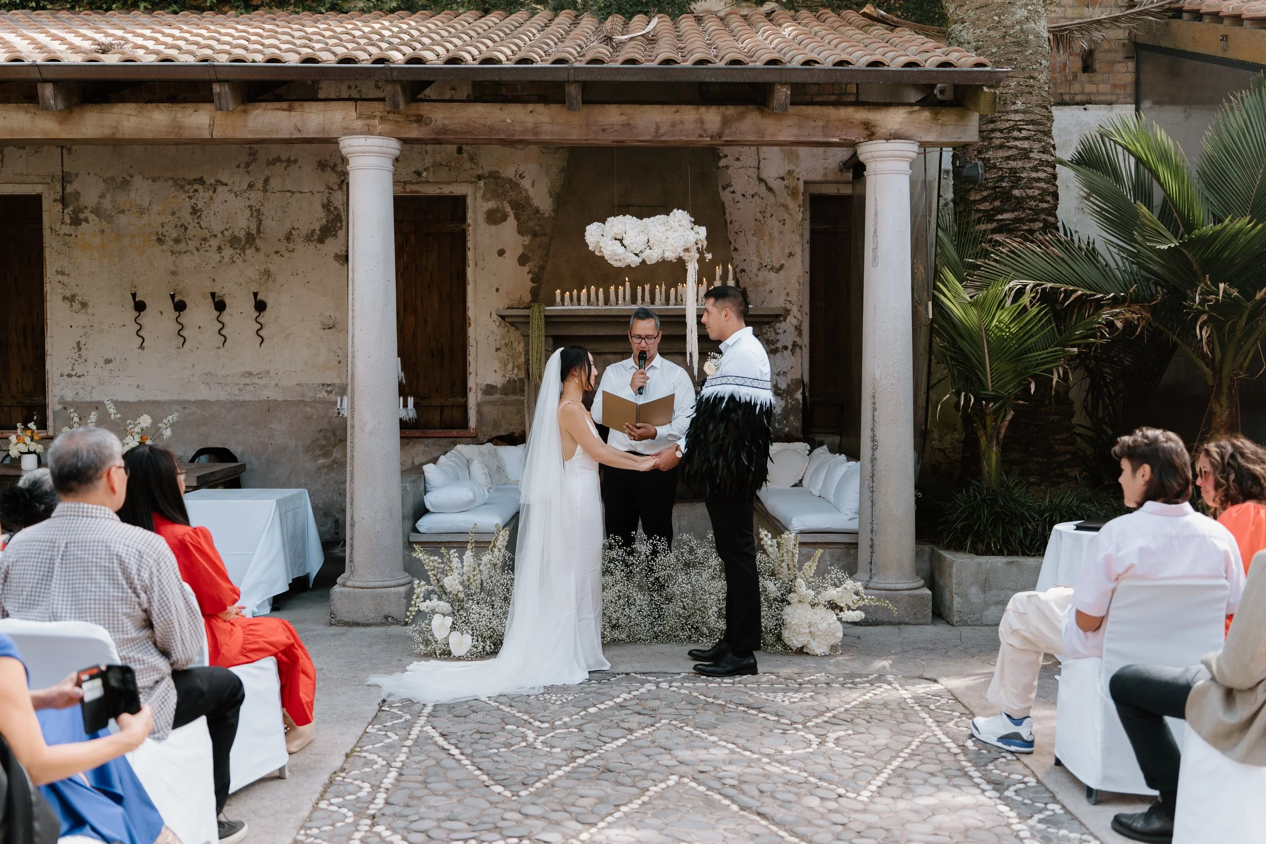 A wedding ceremony taking place outdoors underneath a covered patio with rustic columns and a stone wall background. The bride and groom are holding hands and exchanging vows while the officiant reads from a book. Guests are seated on white chairs on either side, some taking photos. Decor includes white flowers, candles, and a cloud-shaped floral arrangement hanging above.