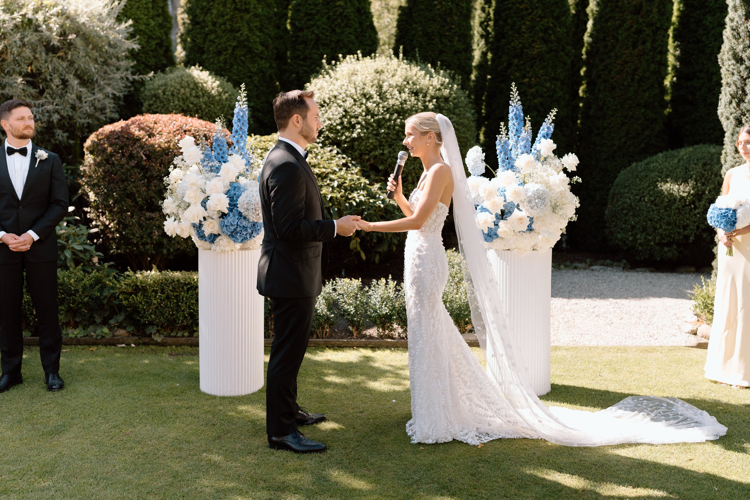 Bride and groom exchanging vows during outdoor wedding ceremony in garden with tall green trees, flowers, and two large white floral arrangements with blue and white flowers in the background.