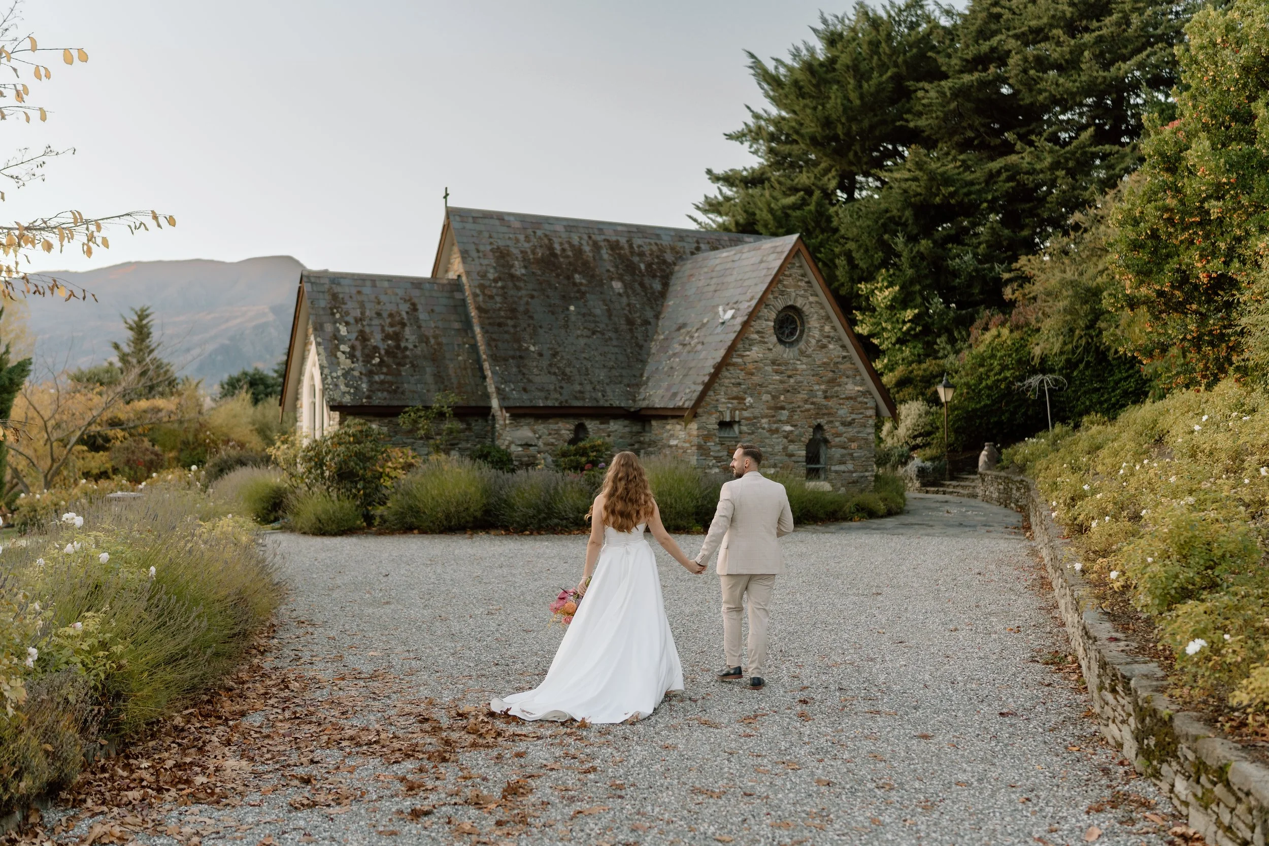 A bride and groom holding hands walk towards a stone church on a gravel path, surrounded by autumn foliage.