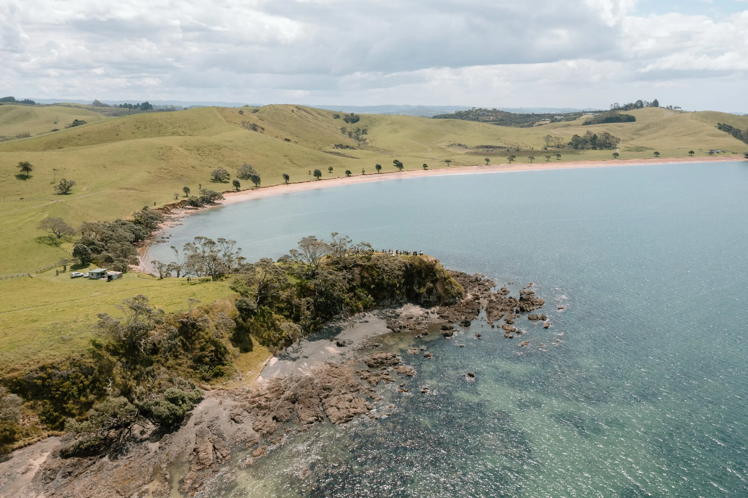 Aerial view of a shoreline with rocky cliffs, sandy beach, and green rolling hills under cloudy sky.