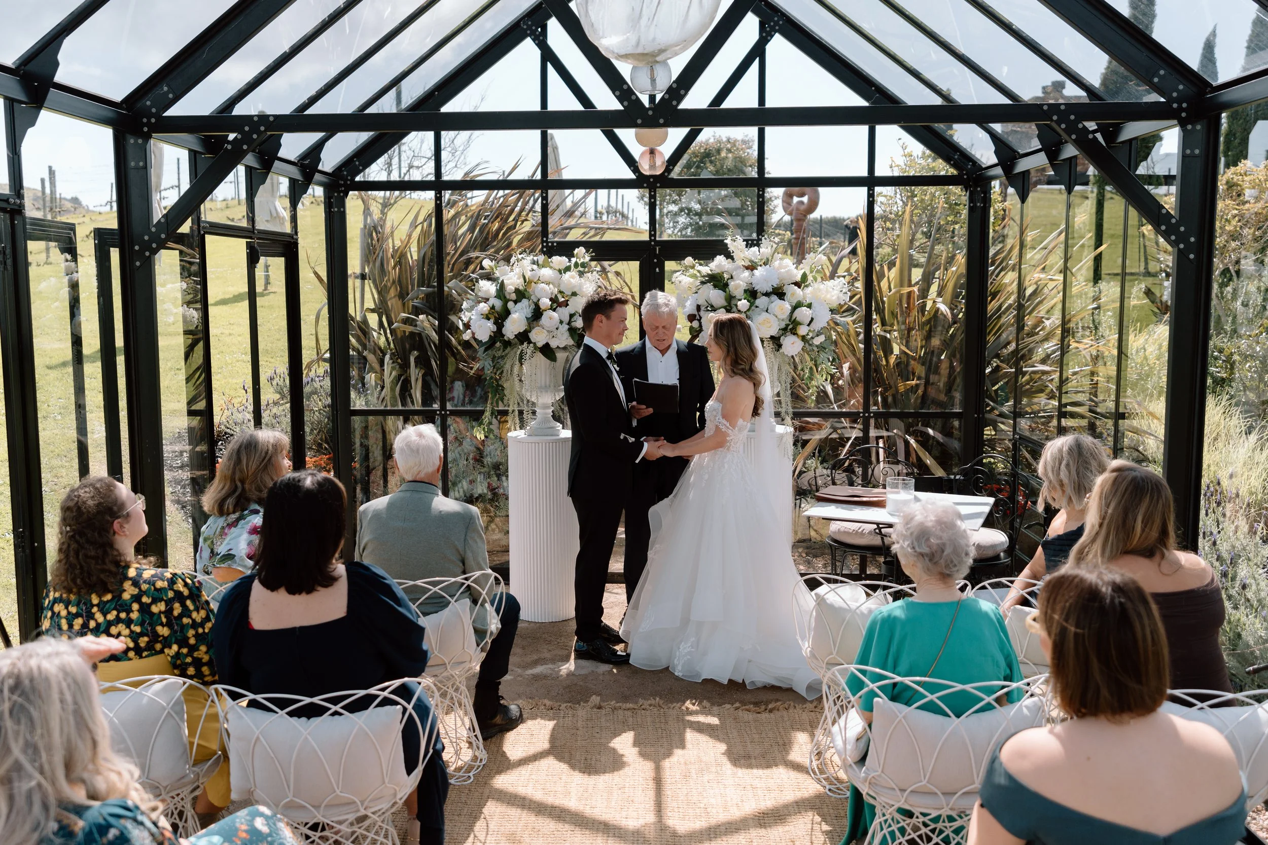 A wedding ceremony taking place inside a glass greenhouse with the bride and groom holding hands and facing each other, an officiant in the center, and guests seated around watching the couple, with large white floral arrangements behind them.