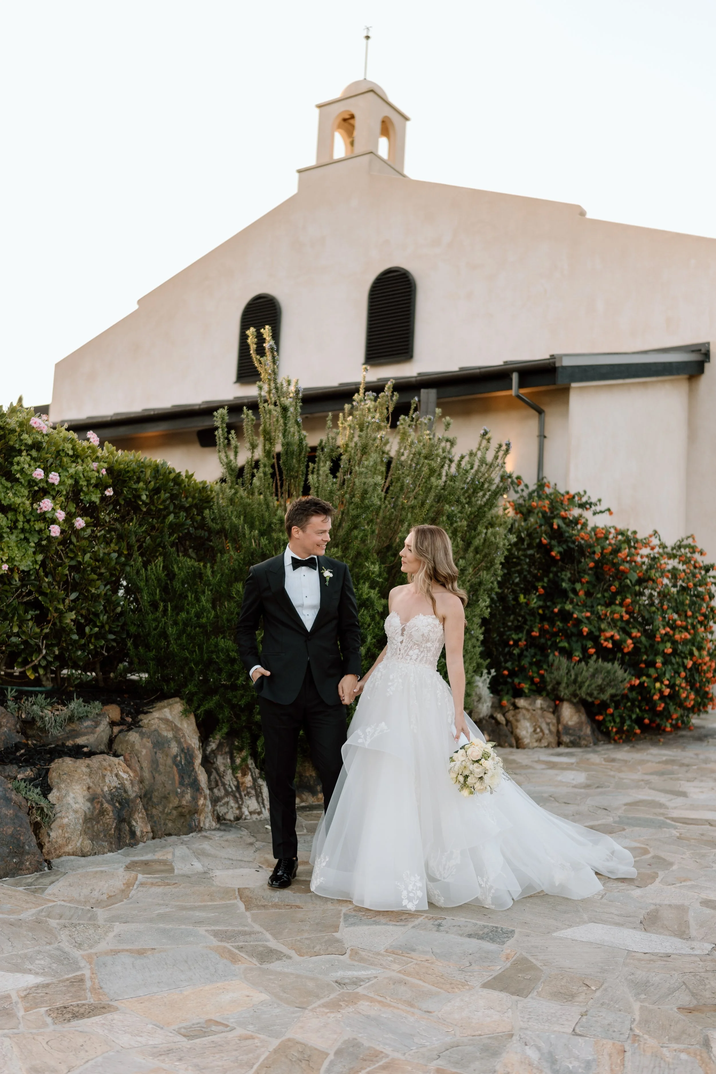 A bride and groom holding hands and smiling at each other outside, with a church building and flowering bushes in the background.