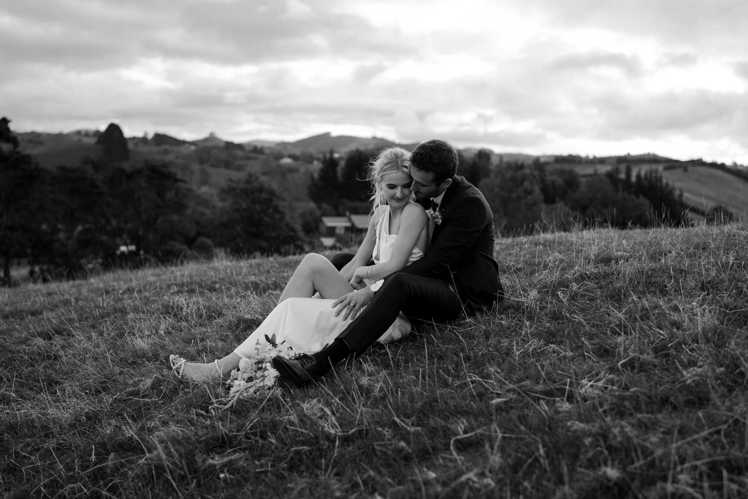 Black and white photo of a newlywed couple sitting on a grassy hill, with the groom whispering in the bride's ear. The bride is smiling with her eyes closed, and the couple is surrounded by a scenic landscape with rolling hills and cloudy sky in the background.