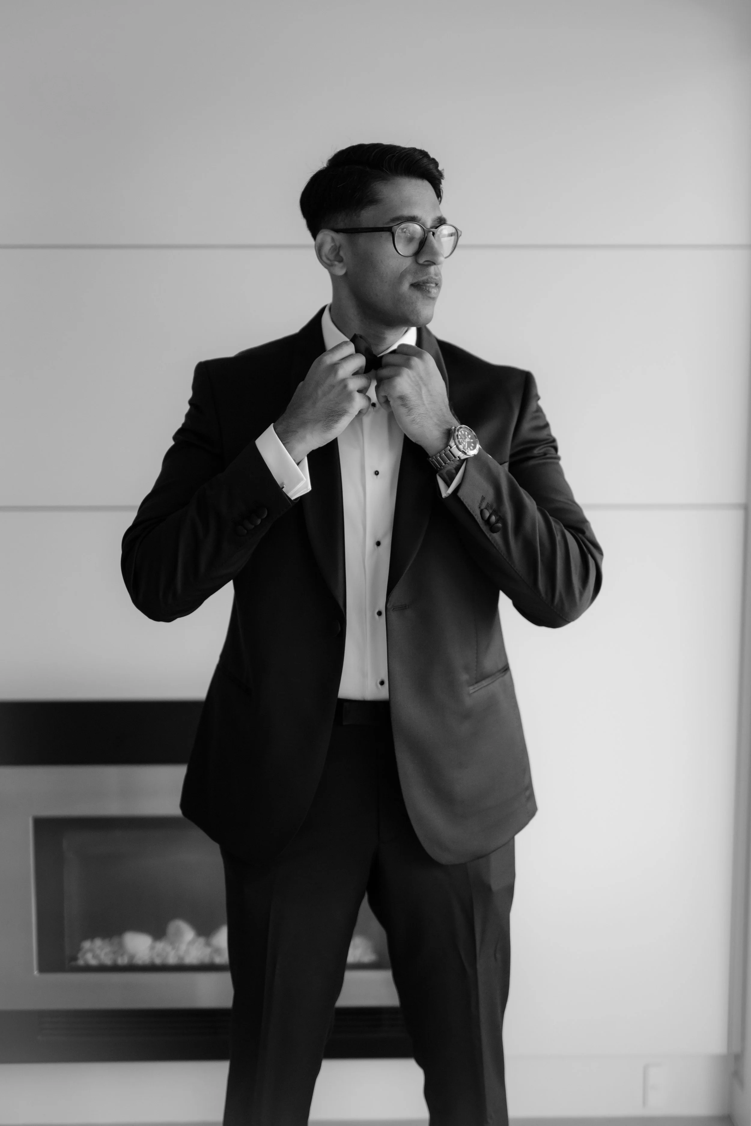 A man in a tuxedo adjusting his bow tie, standing in front of a wall and near a fireplace.