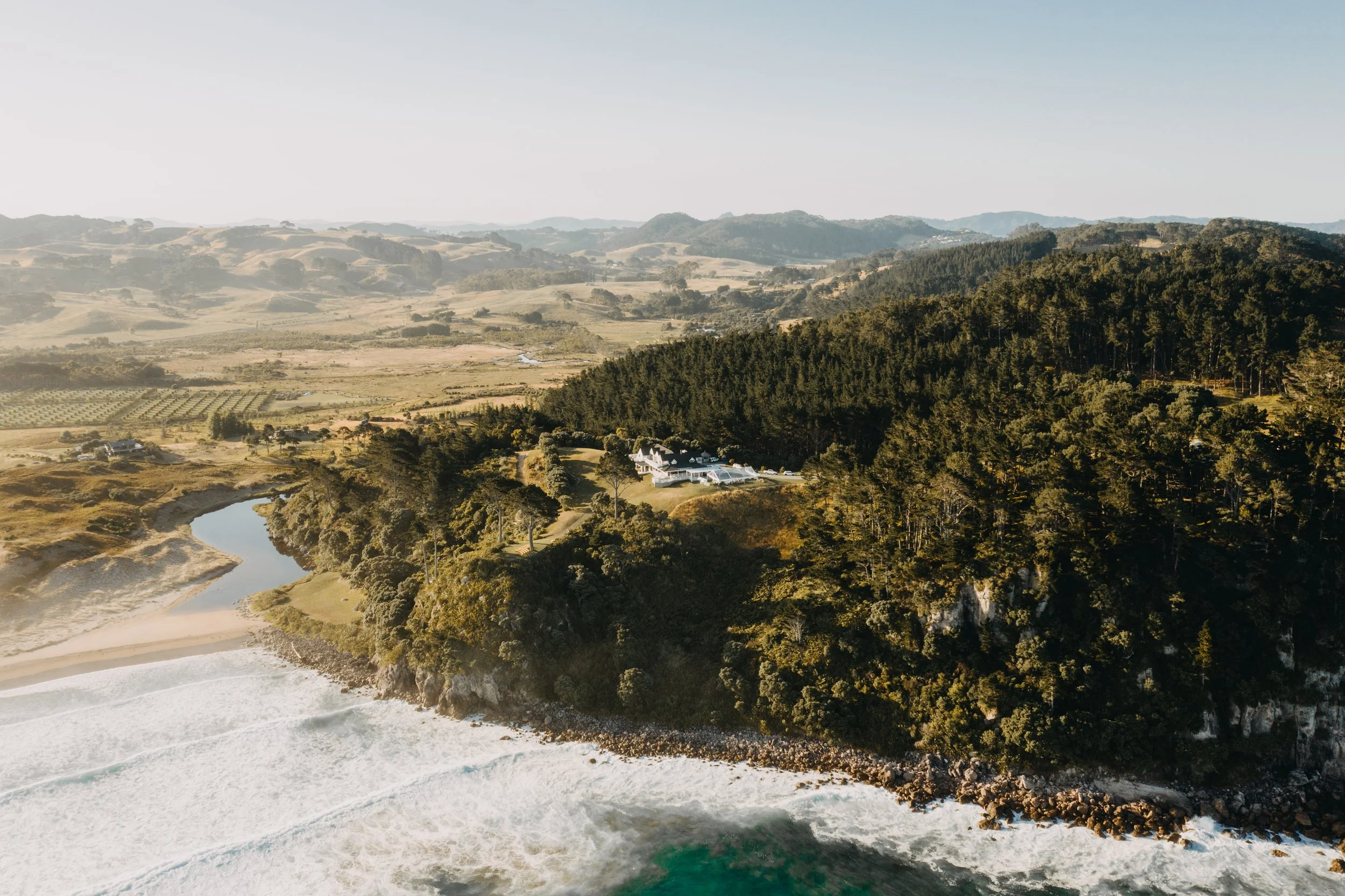 Aerial view of a coastal landscape featuring a forested cliff, a sandy beach, and rolling hills in the background.