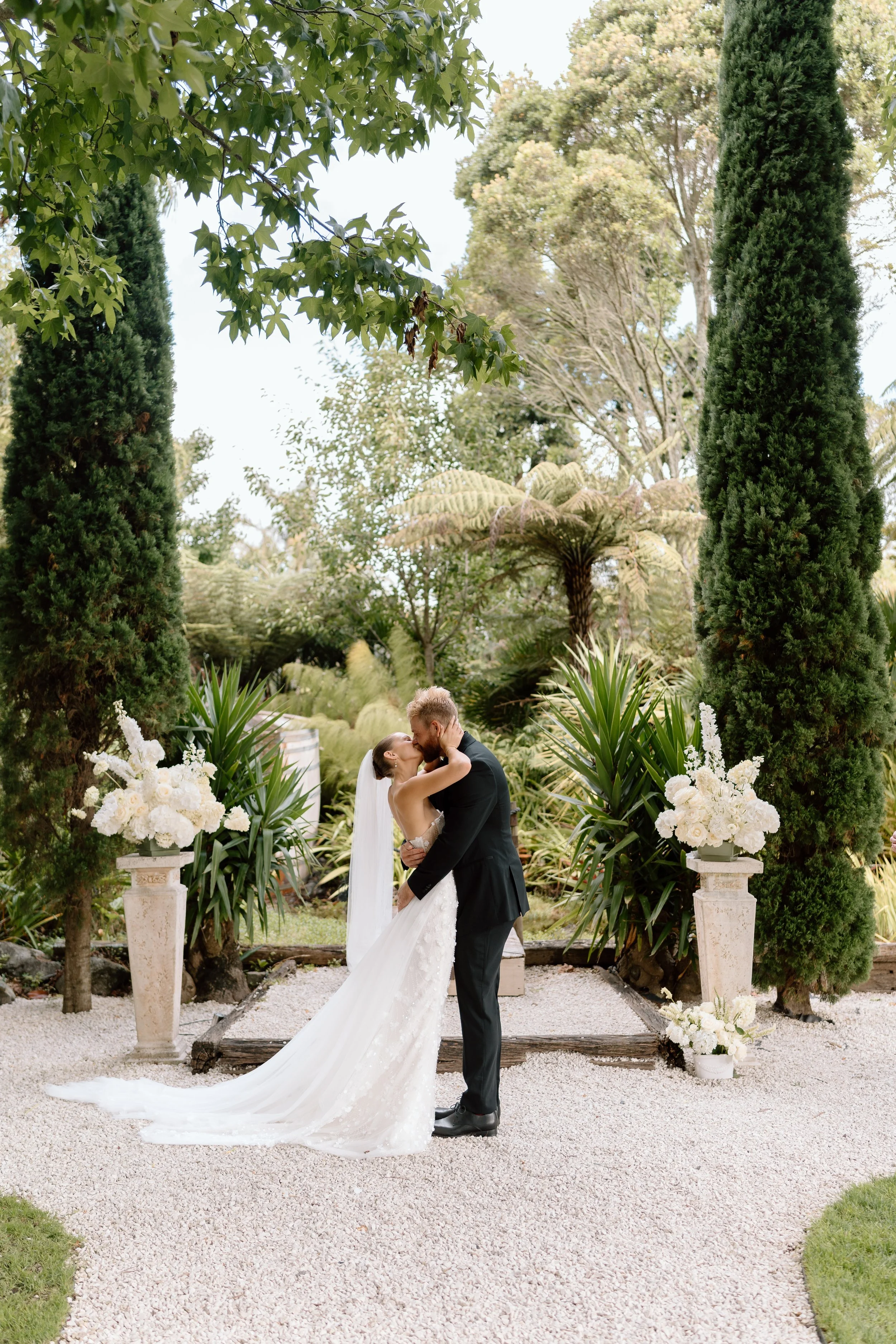 A bride and groom sharing a kiss outdoors, surrounded by tall greenery and white floral arrangements.