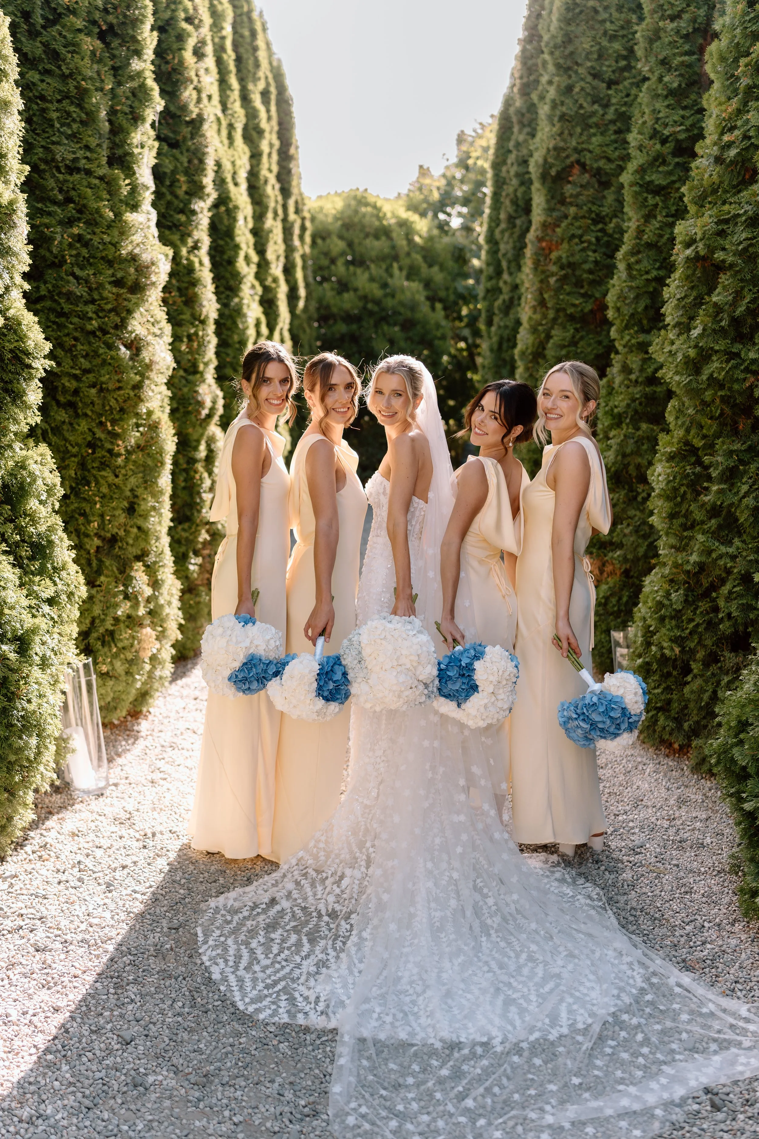 A bride and her bridesmaids holding bouquets stand on a gravel path between tall, green trees under natural sunlight.