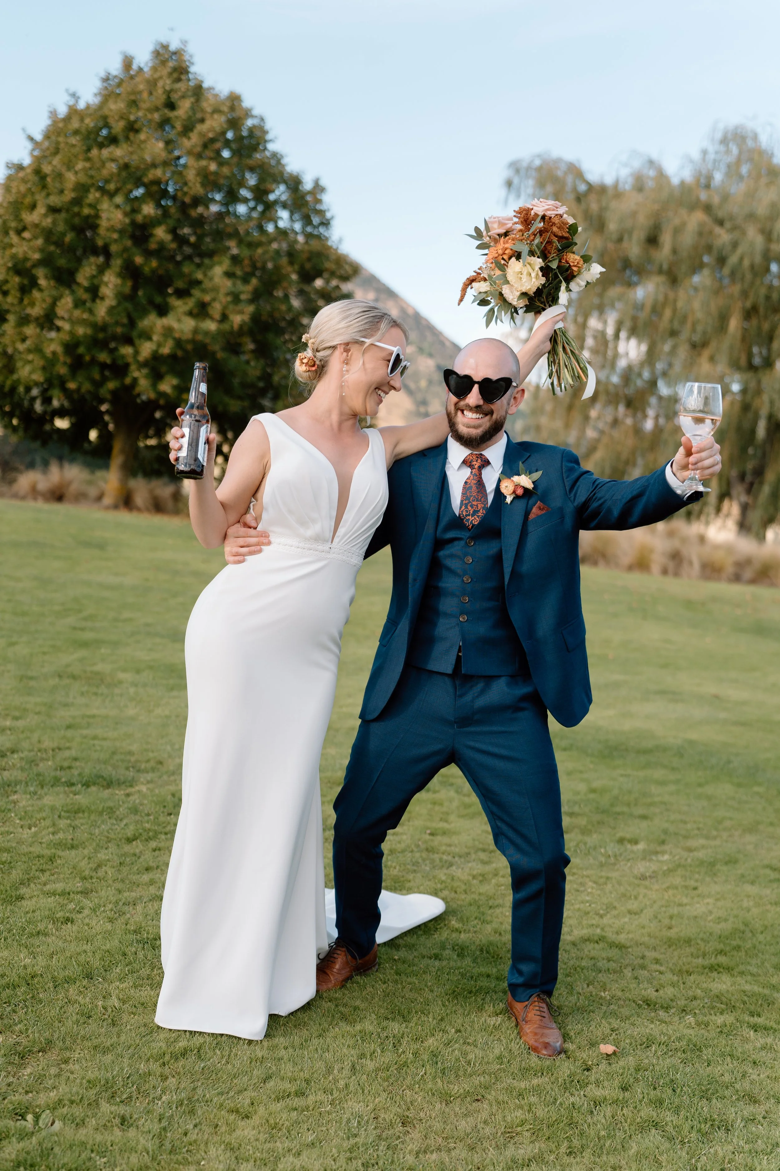 A joyful bride and groom celebrate outdoors on their wedding day, with the bride in a white dress and the groom in a blue suit, both wearing sunglasses, holding drinks, and the bride holding a bouquet of flowers.