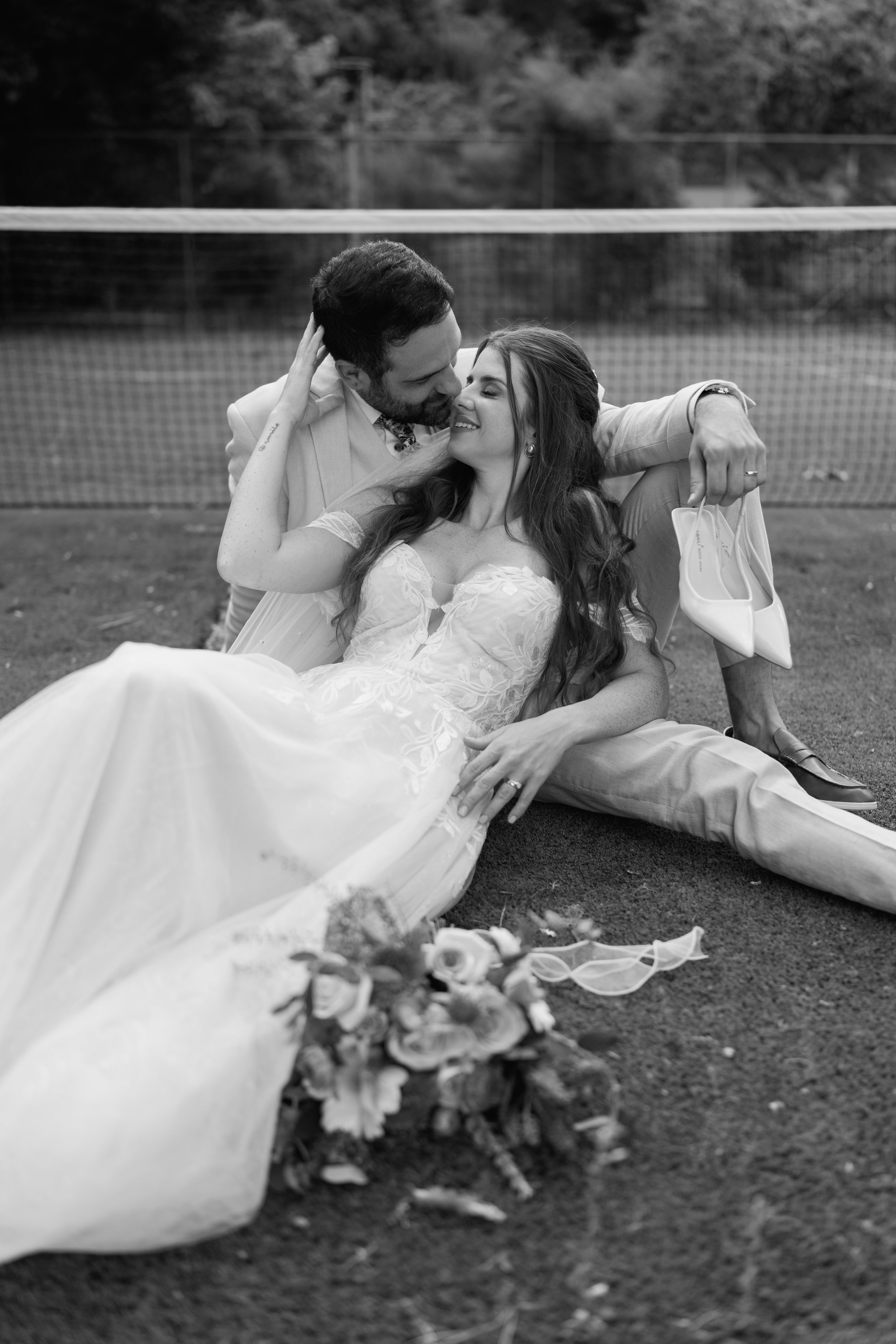 A black and white photo of a bride and groom on a tennis court, with the groom sitting on the ground and the bride lying across his lap. The groom is holding the bride's face and the bride has her hand on the groom's face. There is a bouquet of flowers on the ground near them and some shoes and a face mask nearby.