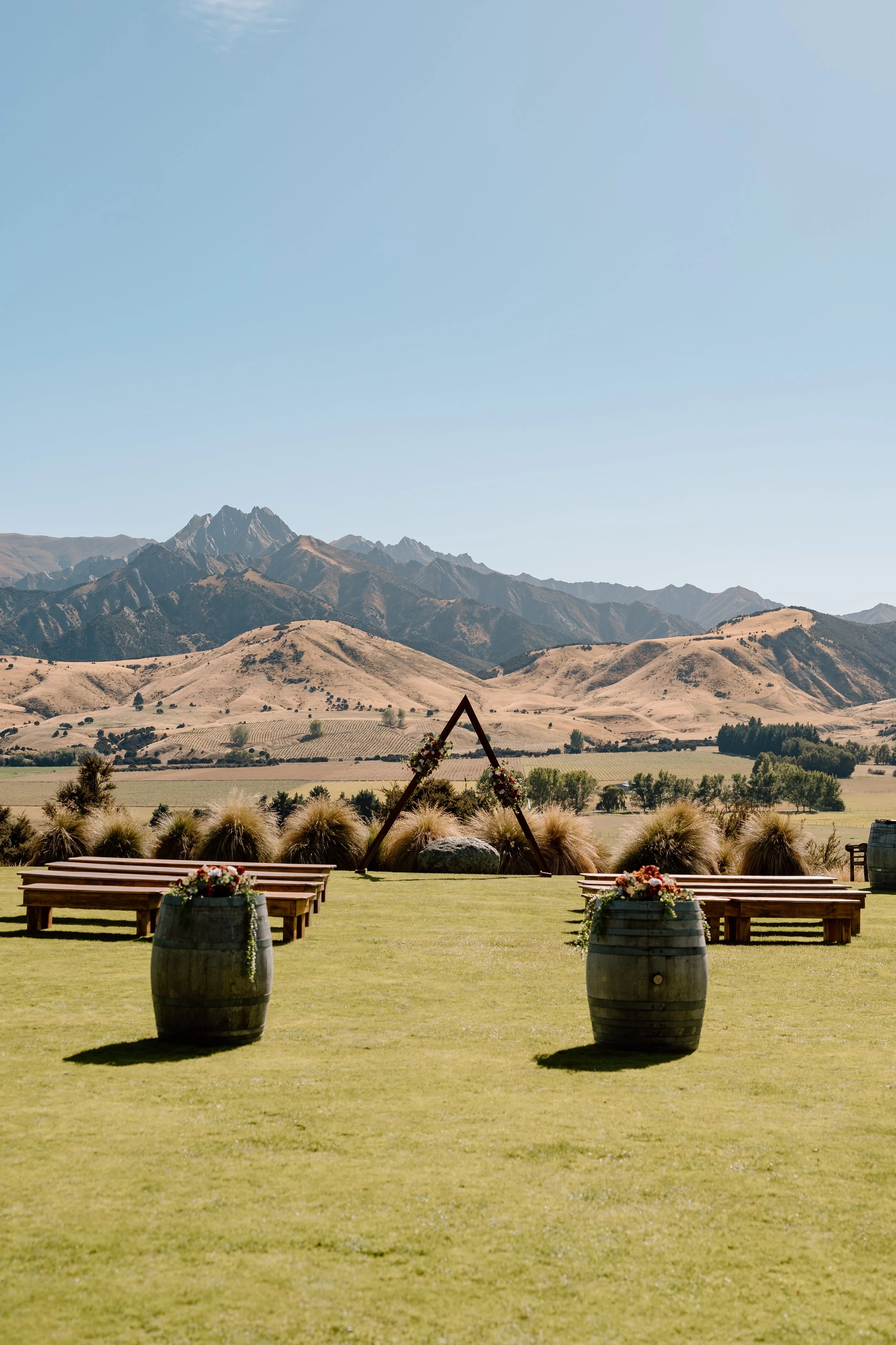 Outdoor wedding setup with wooden benches and barrel planters, scenic mountain landscape in the background, clear blue sky.