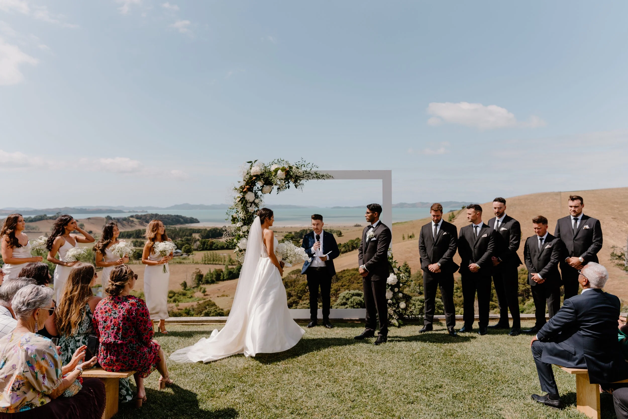 Kauri Bay Outdoor wedding ceremony with bride, groom, officiant, bridesmaids, groomsmen, and guests in scenic landscape with water and hills in the background.