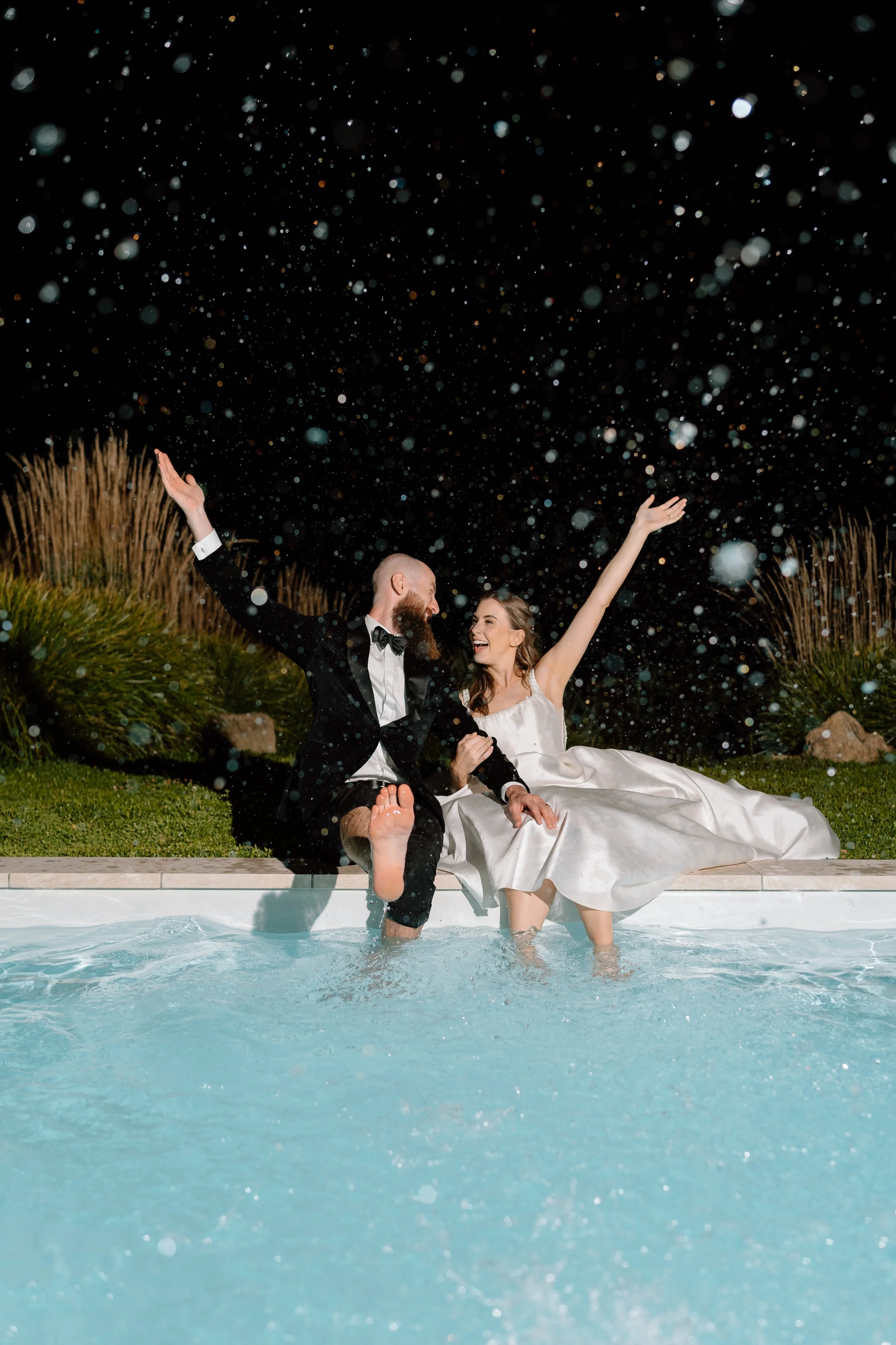 A bride and groom in formal wedding attire sitting by a swimming pool at night, splashing water and celebrating with joyful expressions.