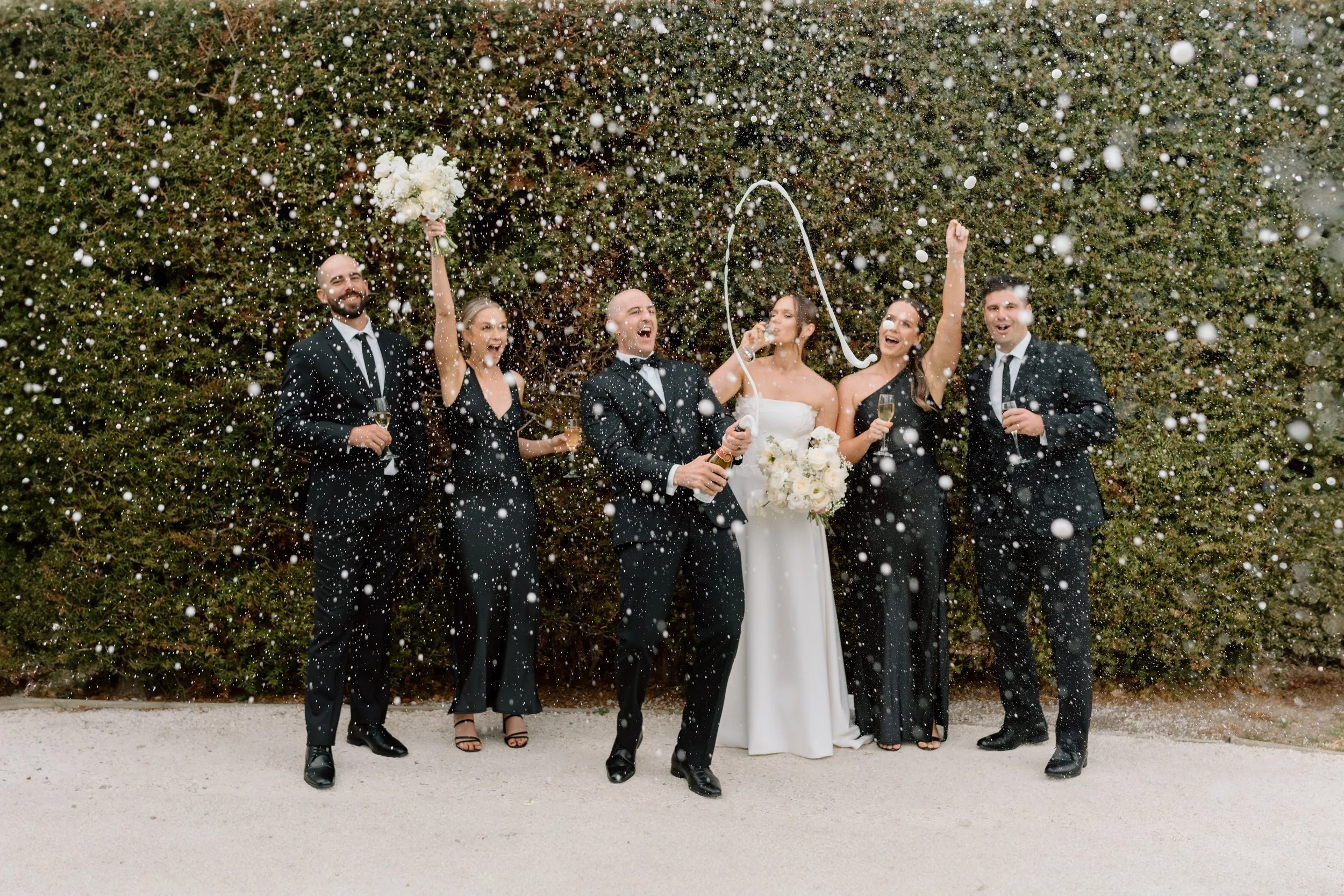 Happy wedding celebration with six people throwing confetti and champagne, outdoors in front of a dense green bush, including a bride in a white dress holding a bouquet, and five guests in formal black suits and dresses.