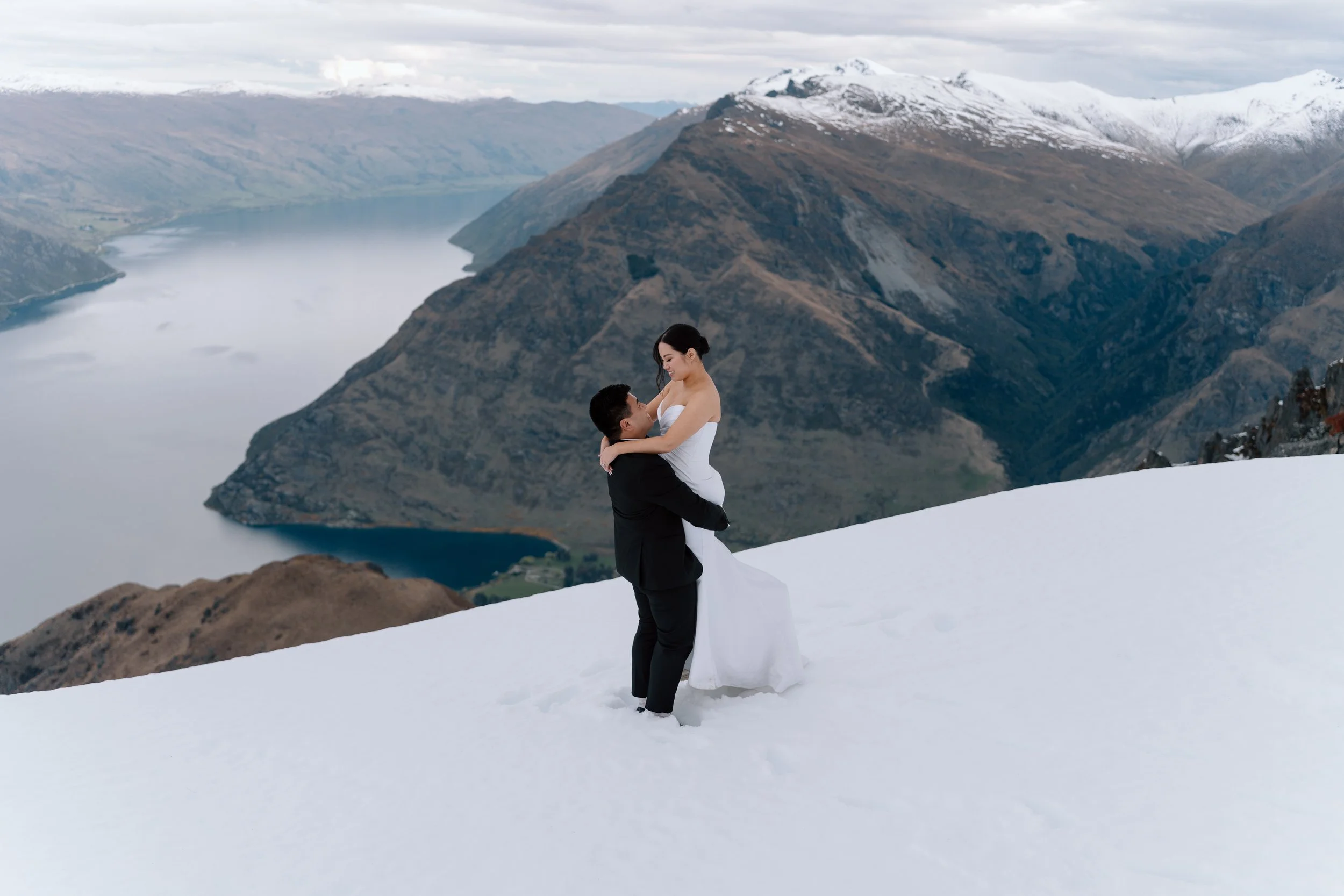 A newlywed couple in wedding attire standing on snow-covered ground in a mountain landscape, with a river and snow-capped mountains in the background.