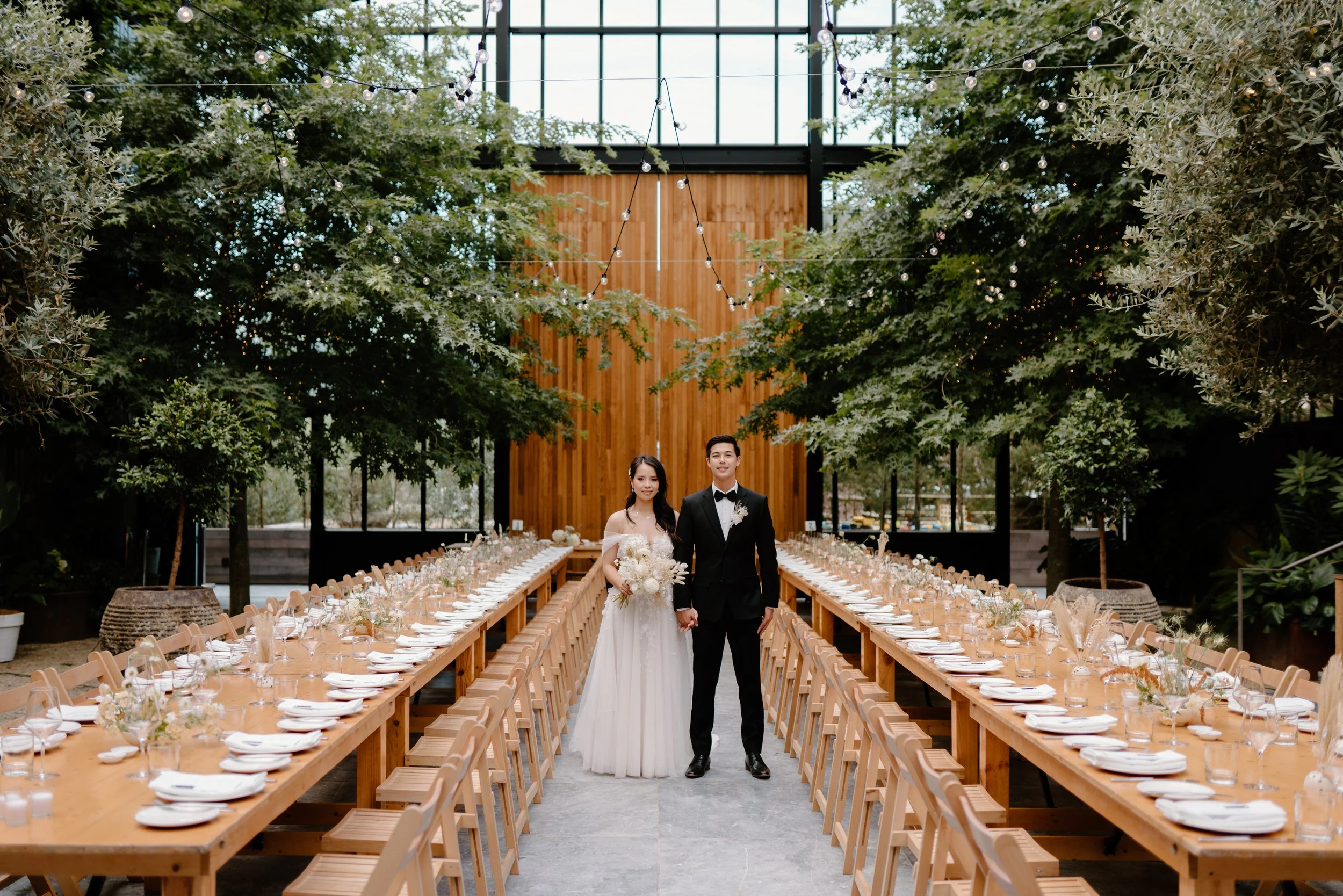 A bride and groom holding hands in the center of a wedding reception venue with long wooden tables on either side, decorated with white plates, glasses, and floral centerpieces, inside a greenhouse-style glass building with greenery and string lights overhead.