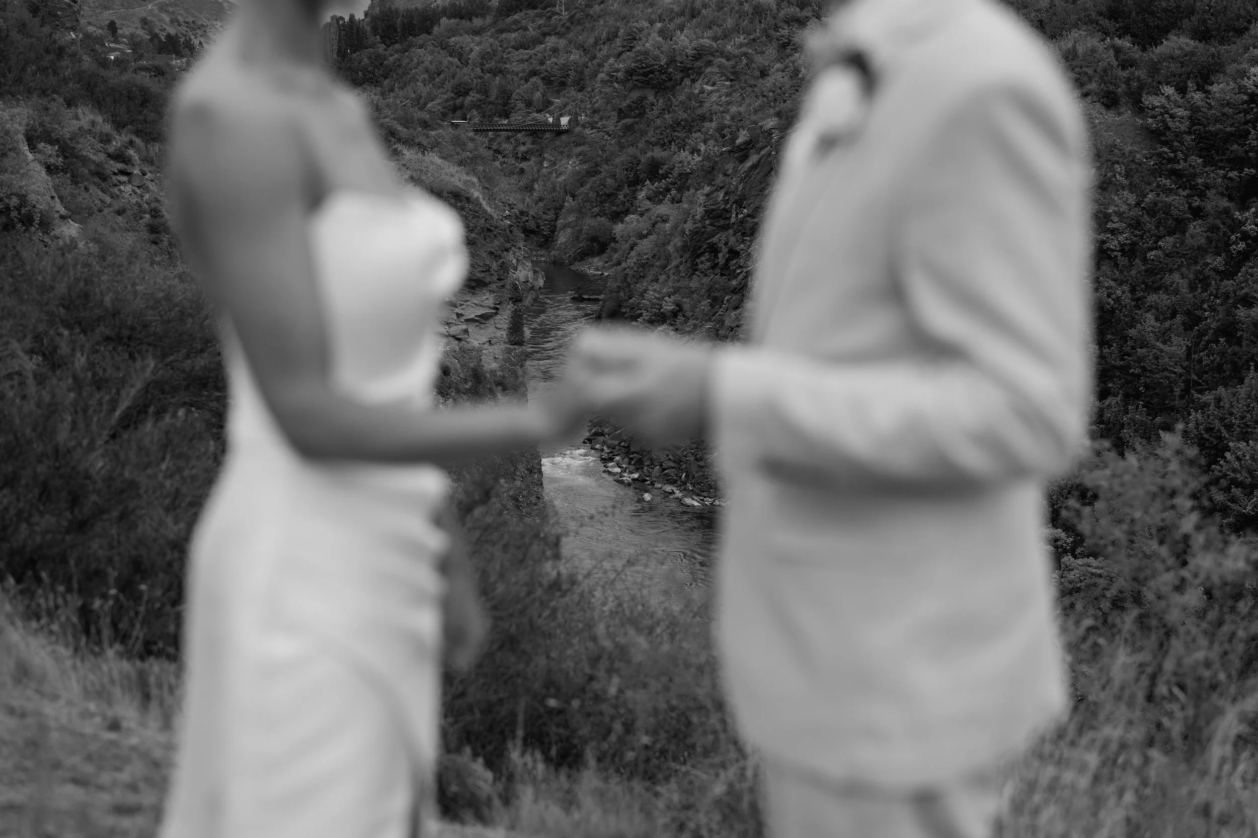 A blurred image of a couple standing outdoors near a river and surrounded by trees, with mountains or hills in the background.