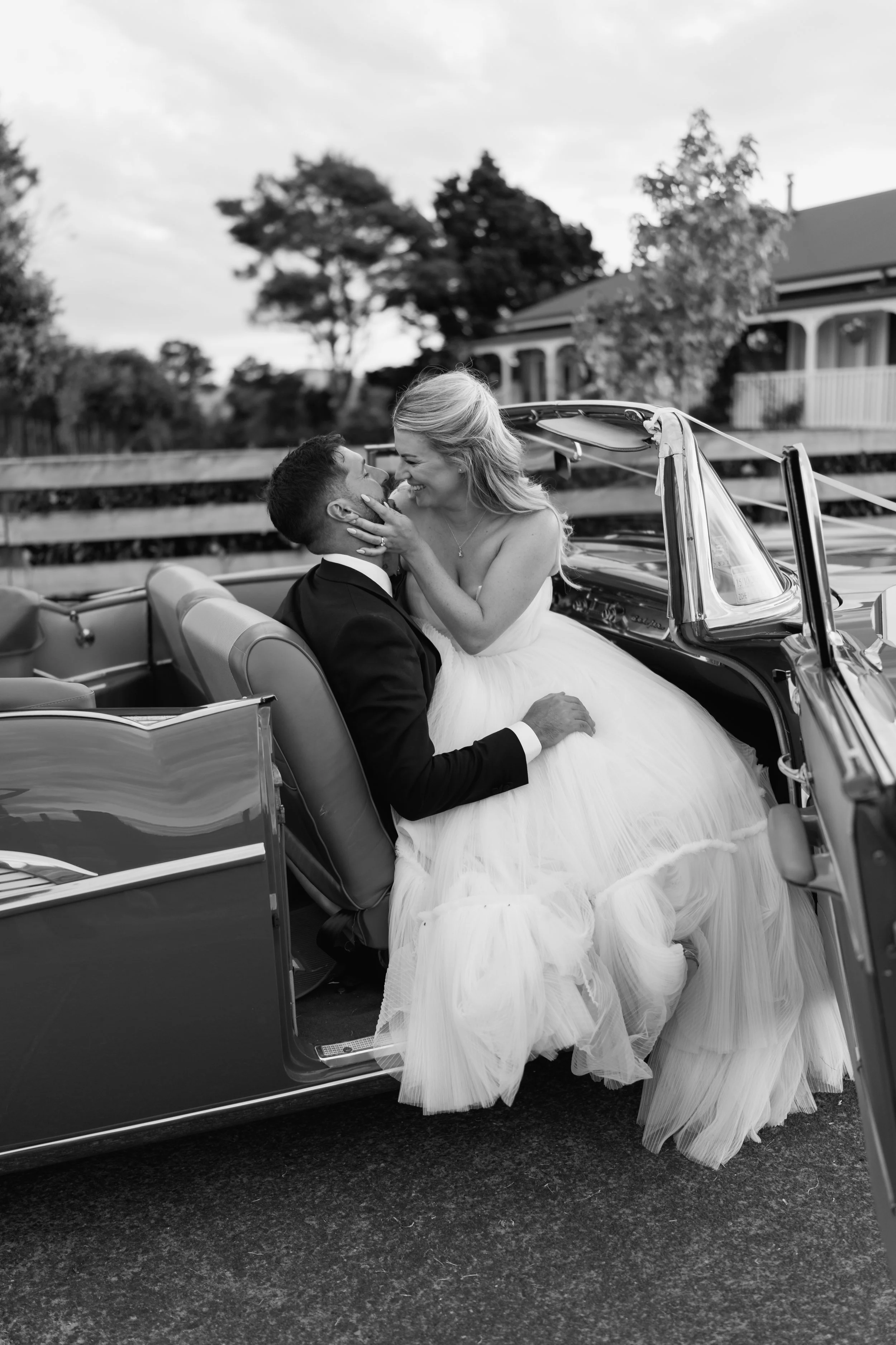 Black and white photo of a bride and groom sitting in a classic convertible car, sharing a romantic moment, with the bride touching the groom's face.