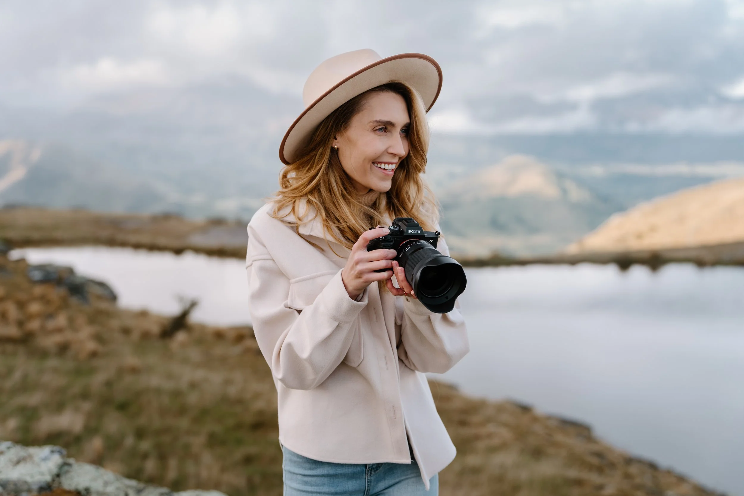 A woman with long blonde hair wearing a beige hat and jacket, holding a Sony camera, standing outdoors near a lake with mountains in the background.
