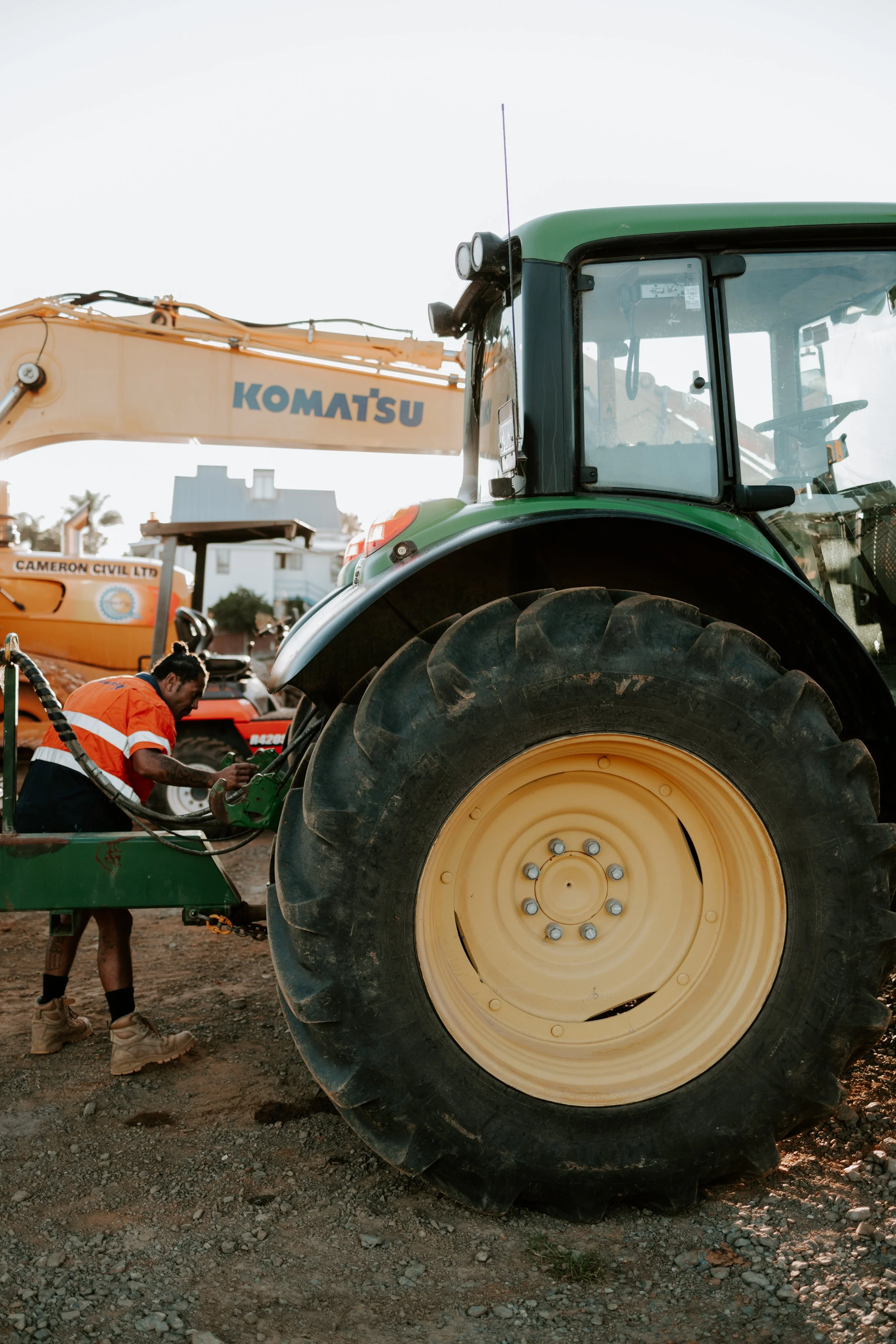 A man working on a large green tractor with yellow wheels in an outdoor area. Another construction vehicle with a Komatsu logo is visible in the background.